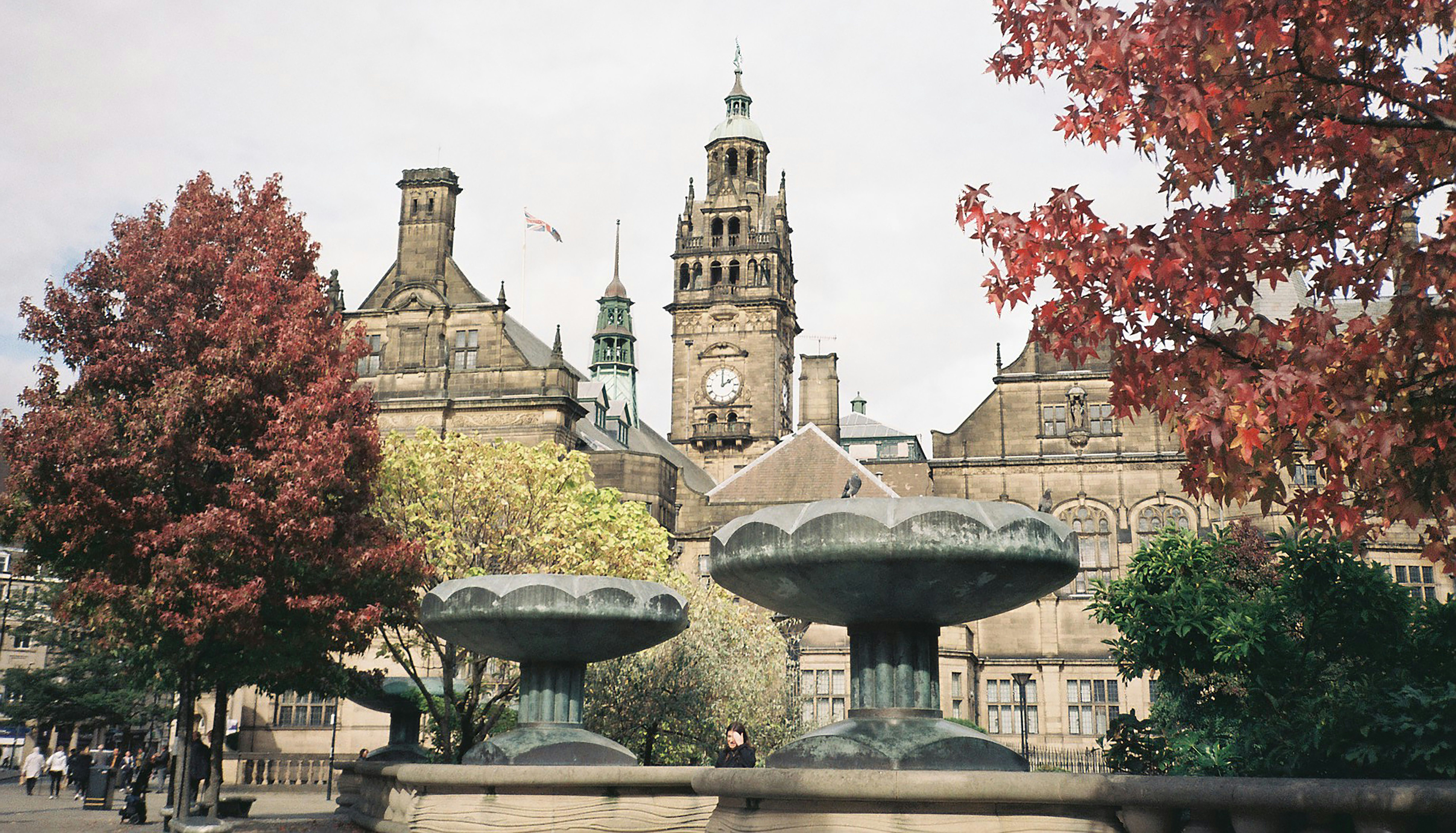 Historic stone building with a clock tower, surrounded by autumn trees and large ornate fountains in the foreground.