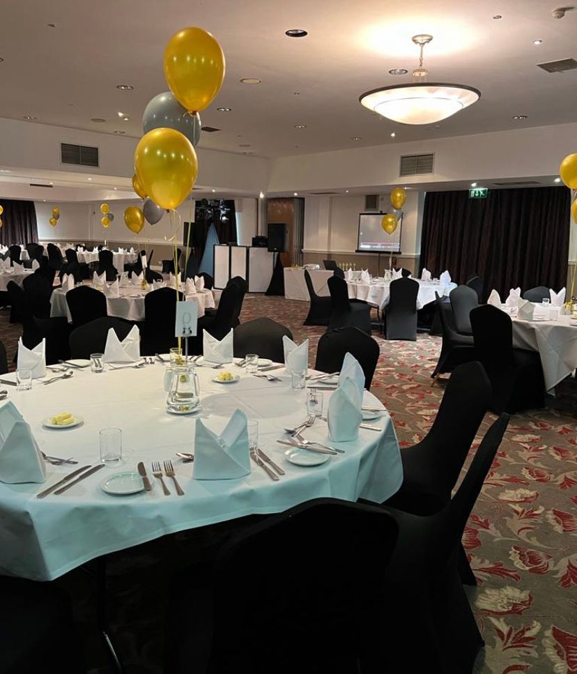 Banquet hall set up with round tables, black chairs, white tablecloths, and gold balloons.