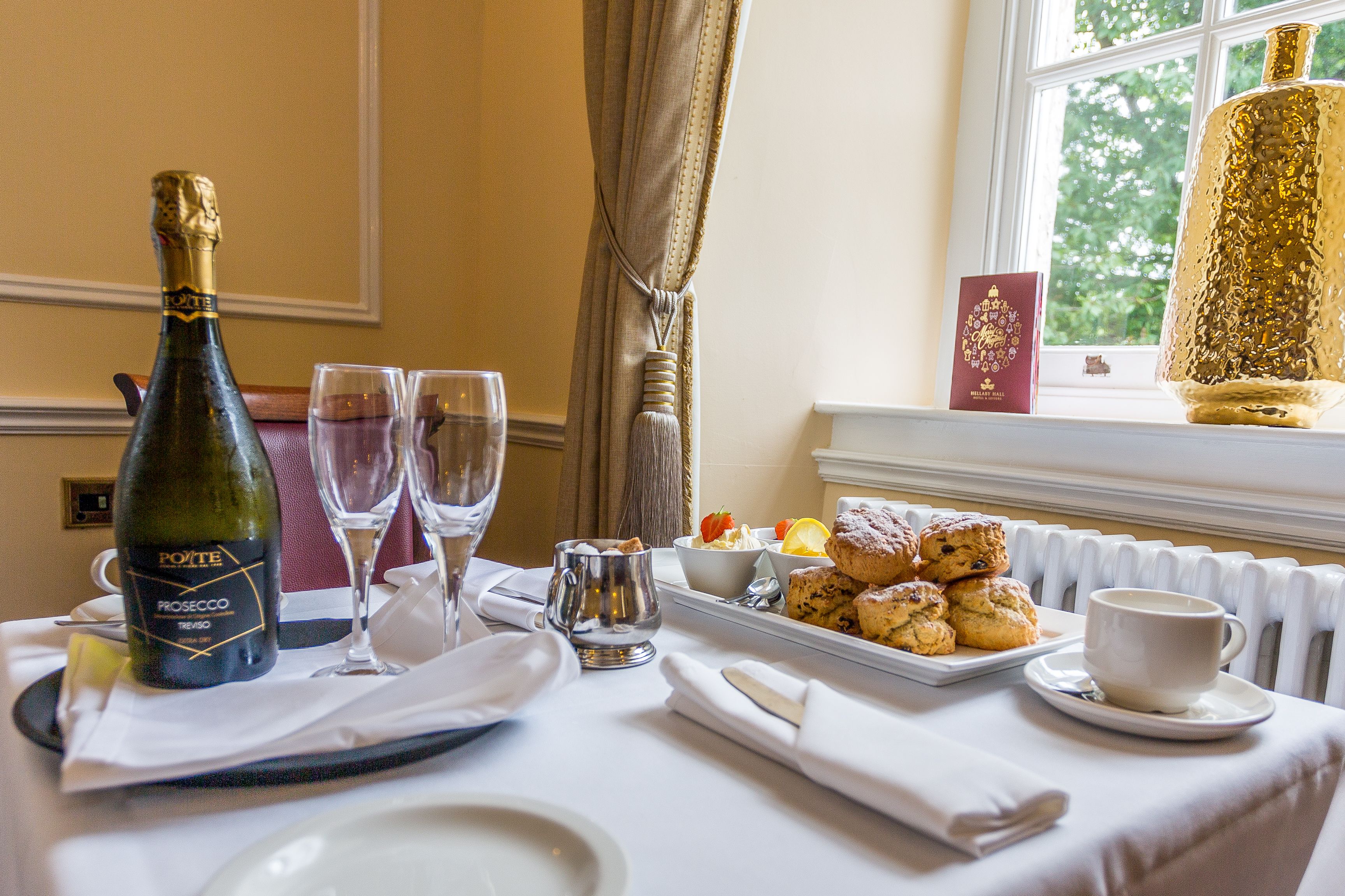 A table set for afternoon tea with a bottle of prosecco, two glasses, scones, a cup of tea, and condiments by a window.