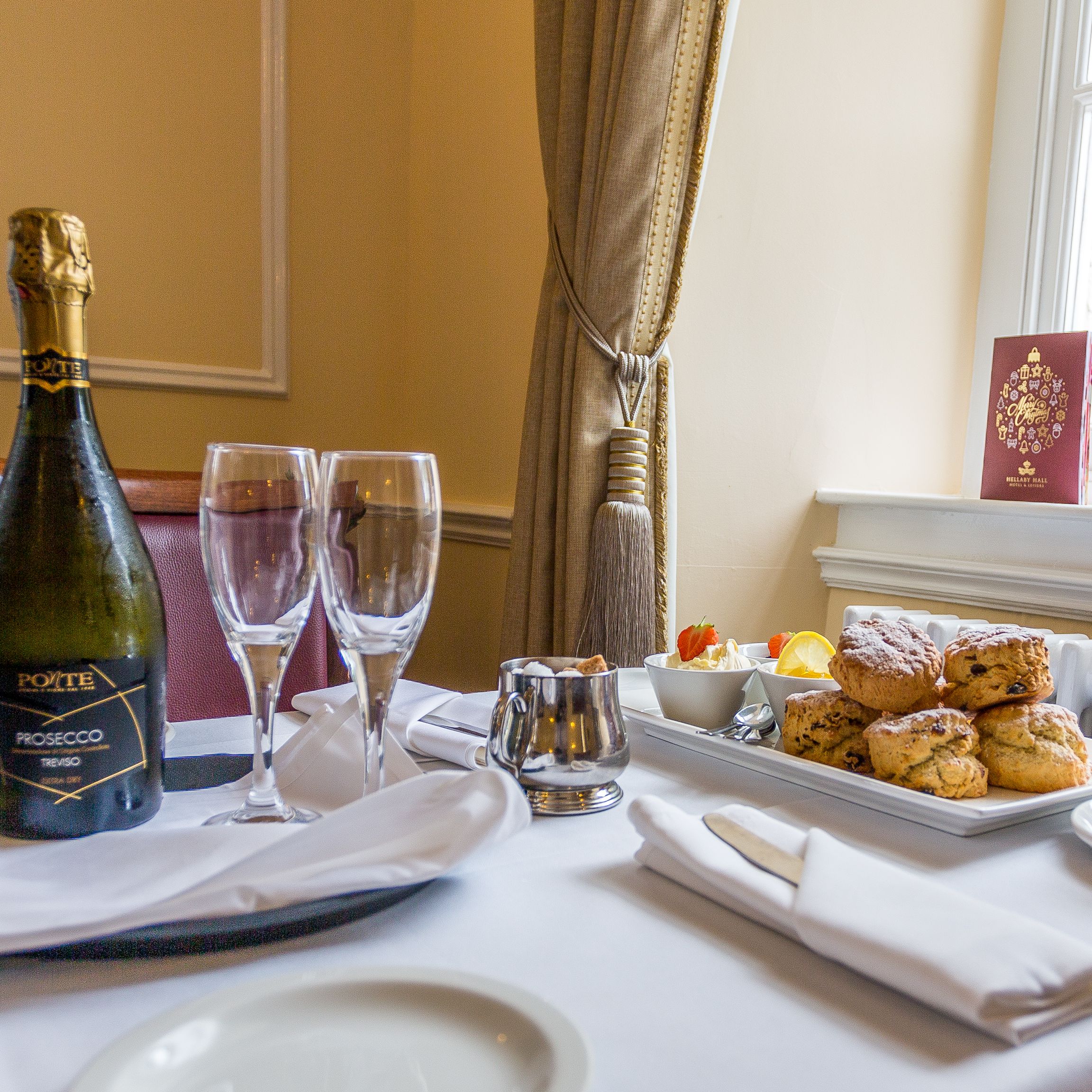 A table set for afternoon tea with a bottle of prosecco, two glasses, scones, a cup of tea, and condiments by a window.
