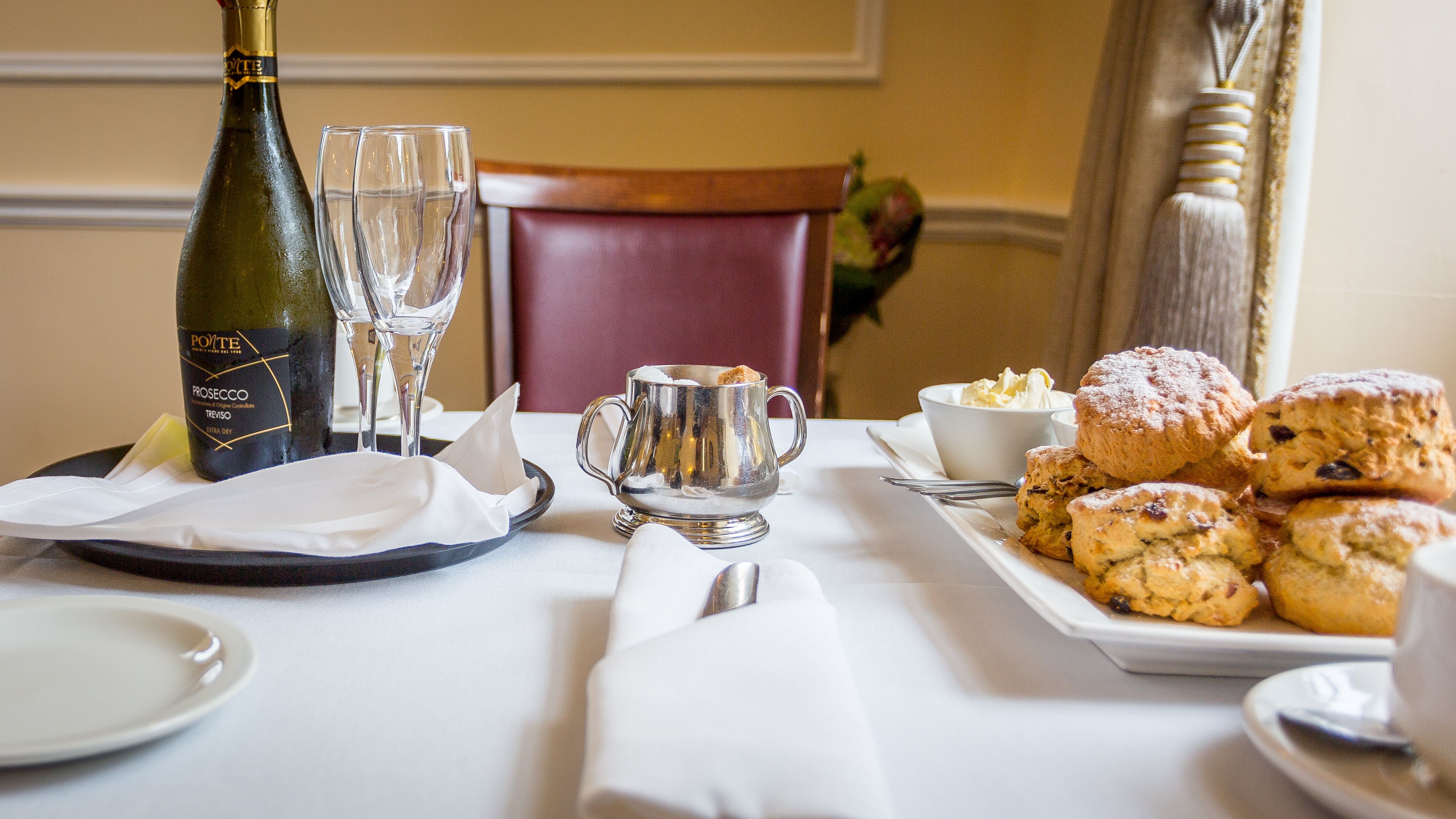 Table set for afternoon tea with a bottle of prosecco, glasses, scones, clotted cream, and silverware.