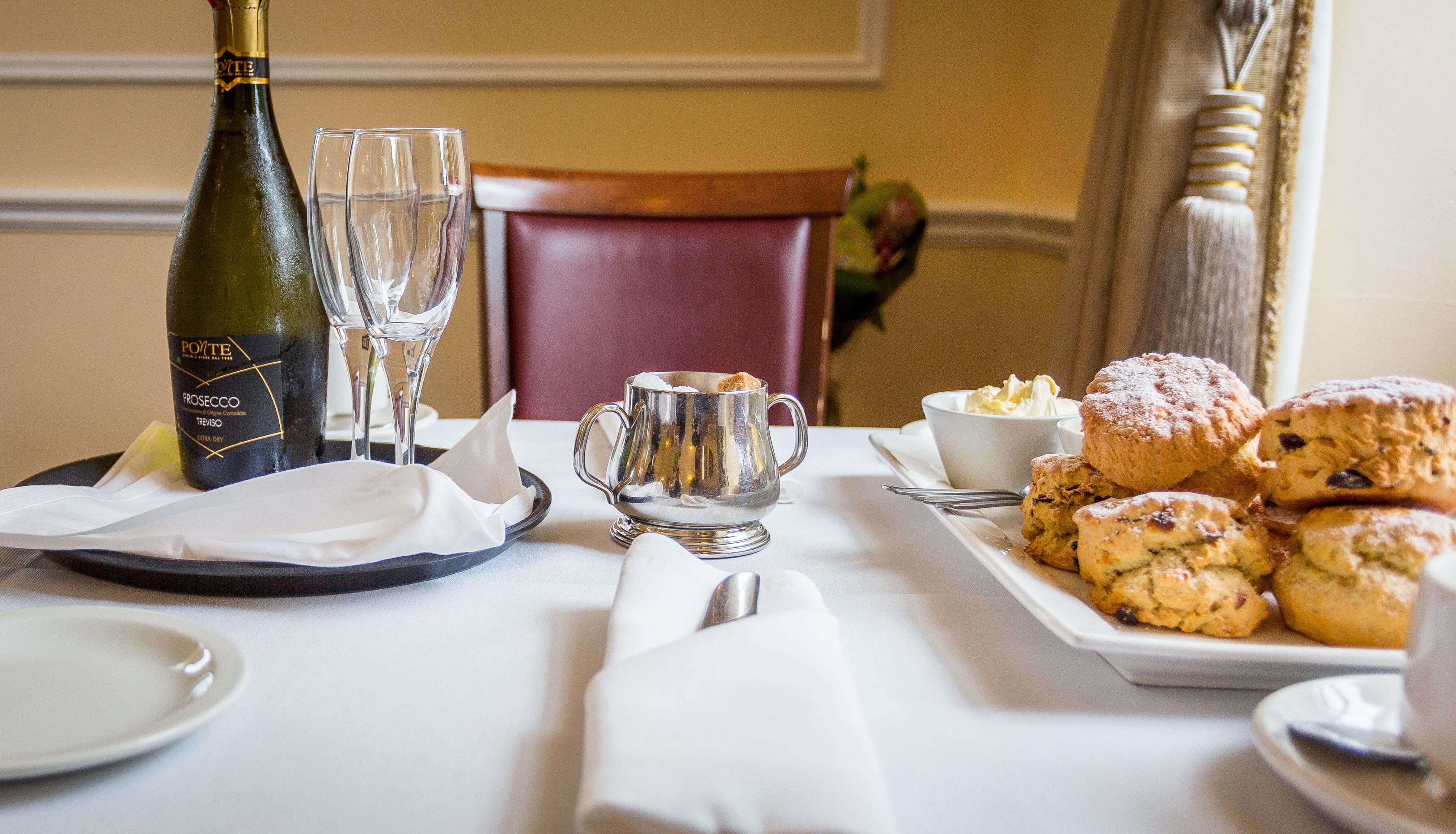 Table set for afternoon tea with a bottle of prosecco, glasses, scones, clotted cream, and silverware.