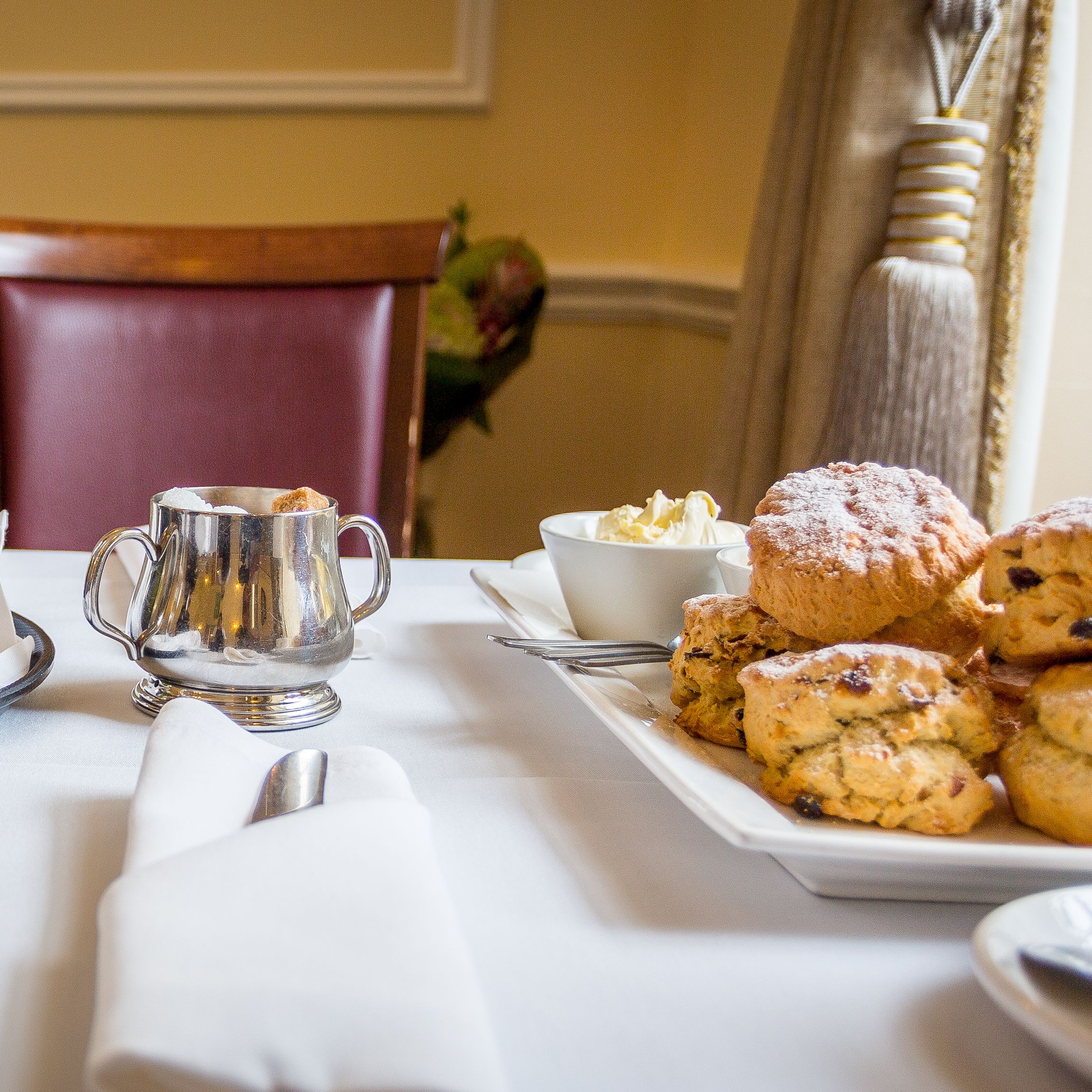 Table set for afternoon tea with a bottle of prosecco, glasses, scones, clotted cream, and silverware.