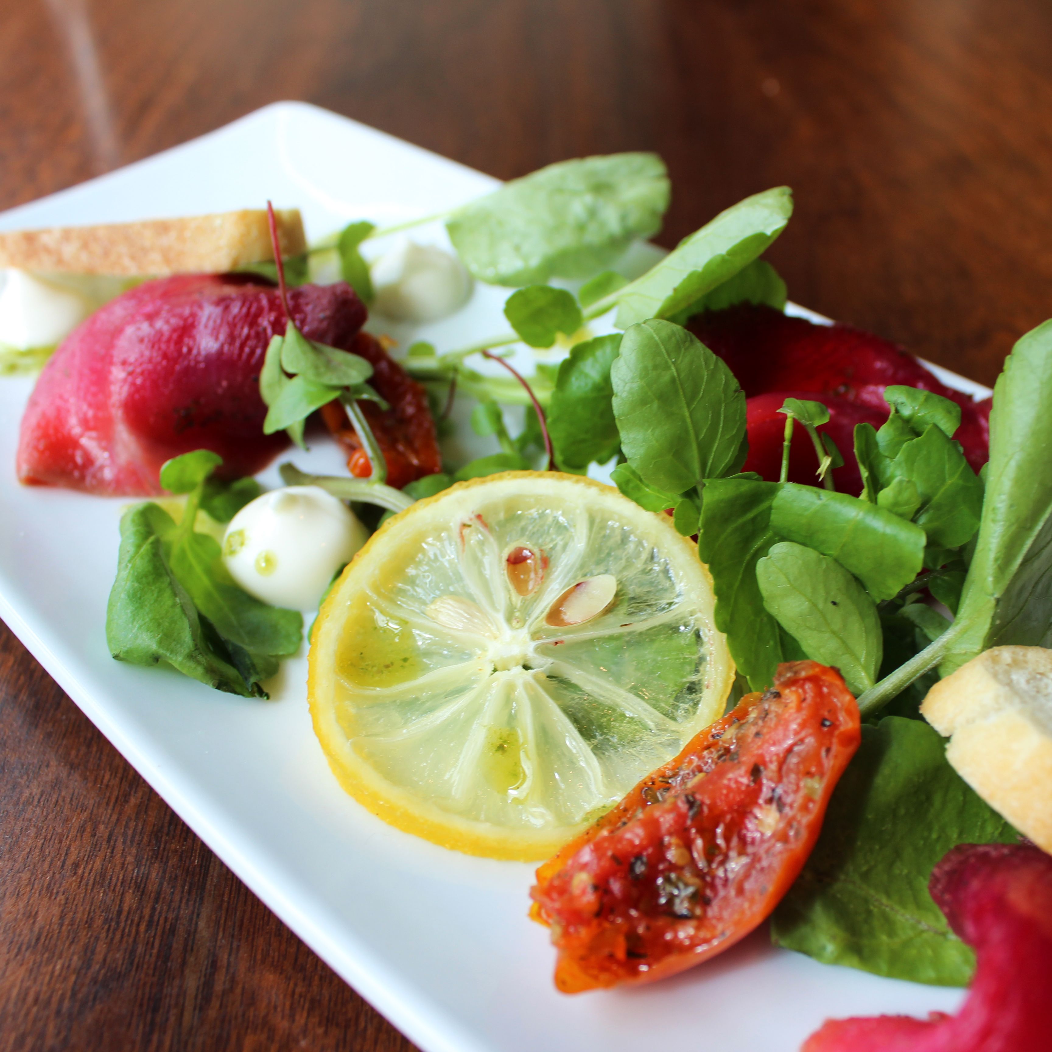 A plated dish featuring greens, a lemon slice, roasted tomato, thin bread slices, and beetroot with dollops of creamy sauce.