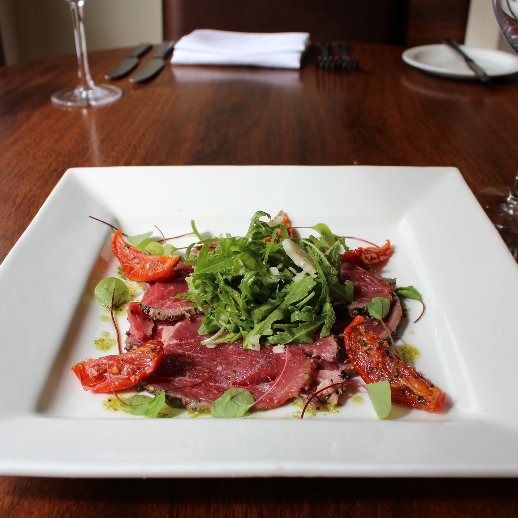 A plate of carpaccio with arugula and roasted tomatoes served on a square white plate