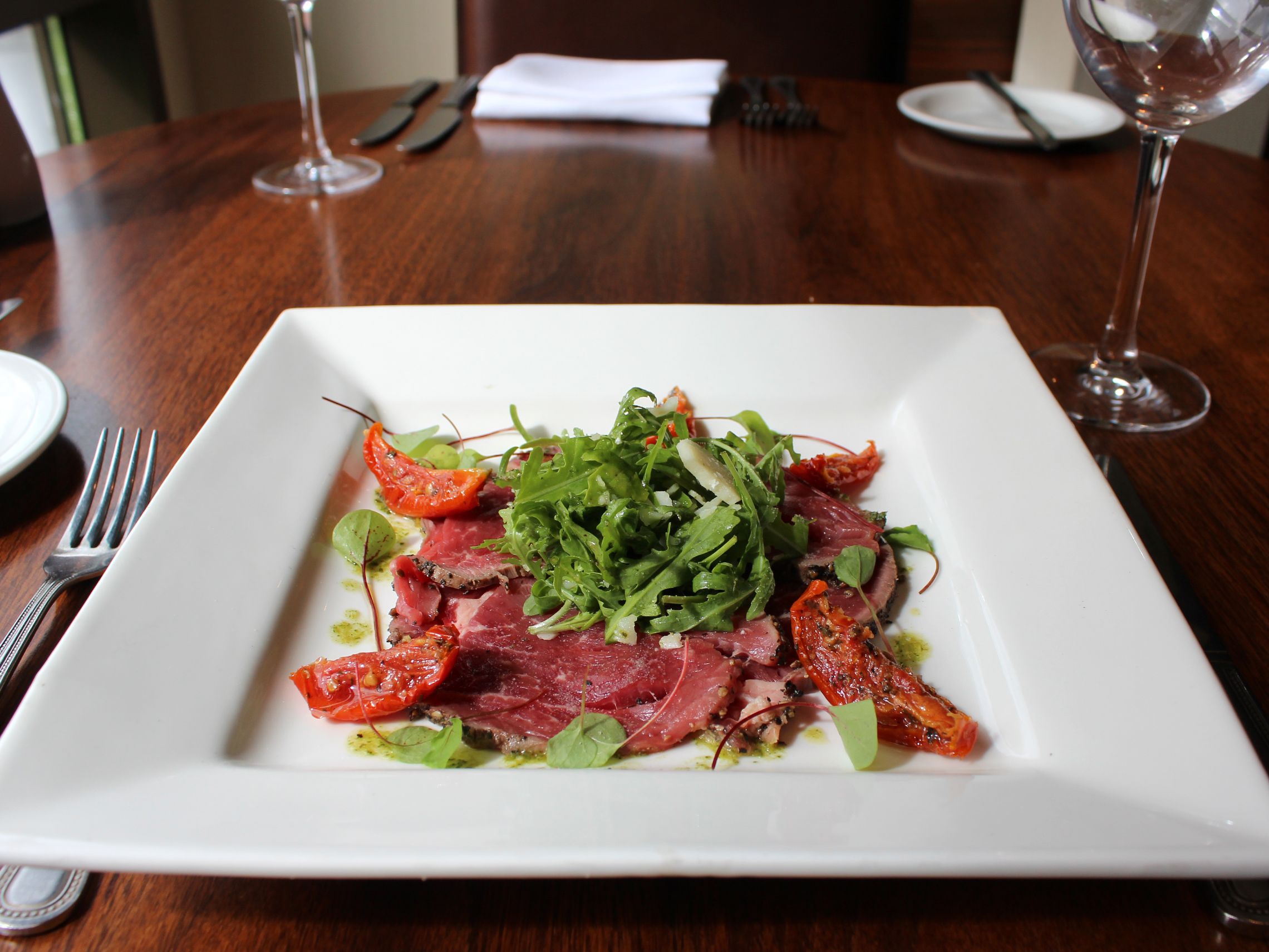 A plate of carpaccio with arugula and roasted tomatoes served on a square white plate