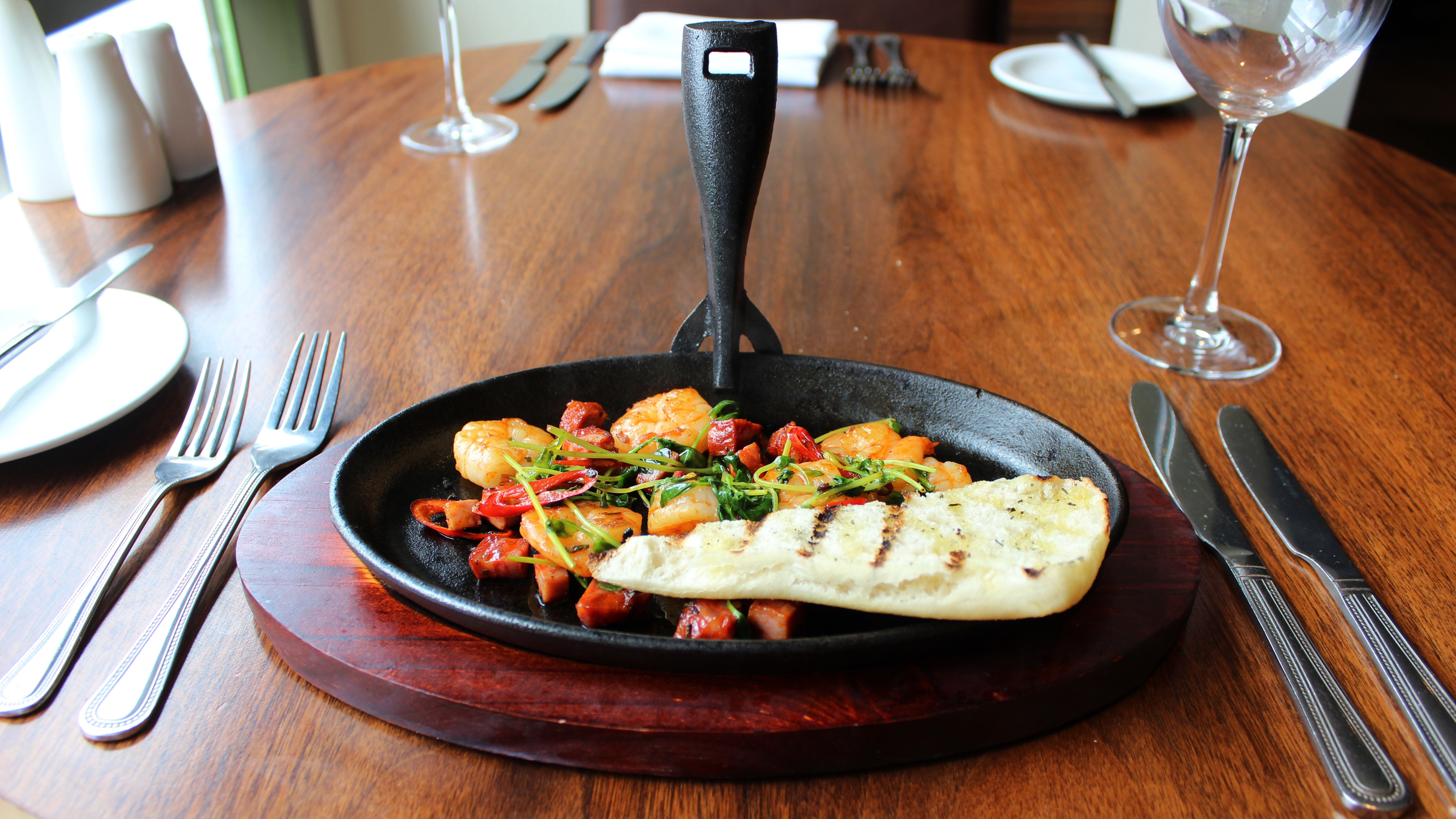 A sizzling skillet of shrimp, diced sausage, vegetables, and grilled bread on a wooden table at a restaurant.