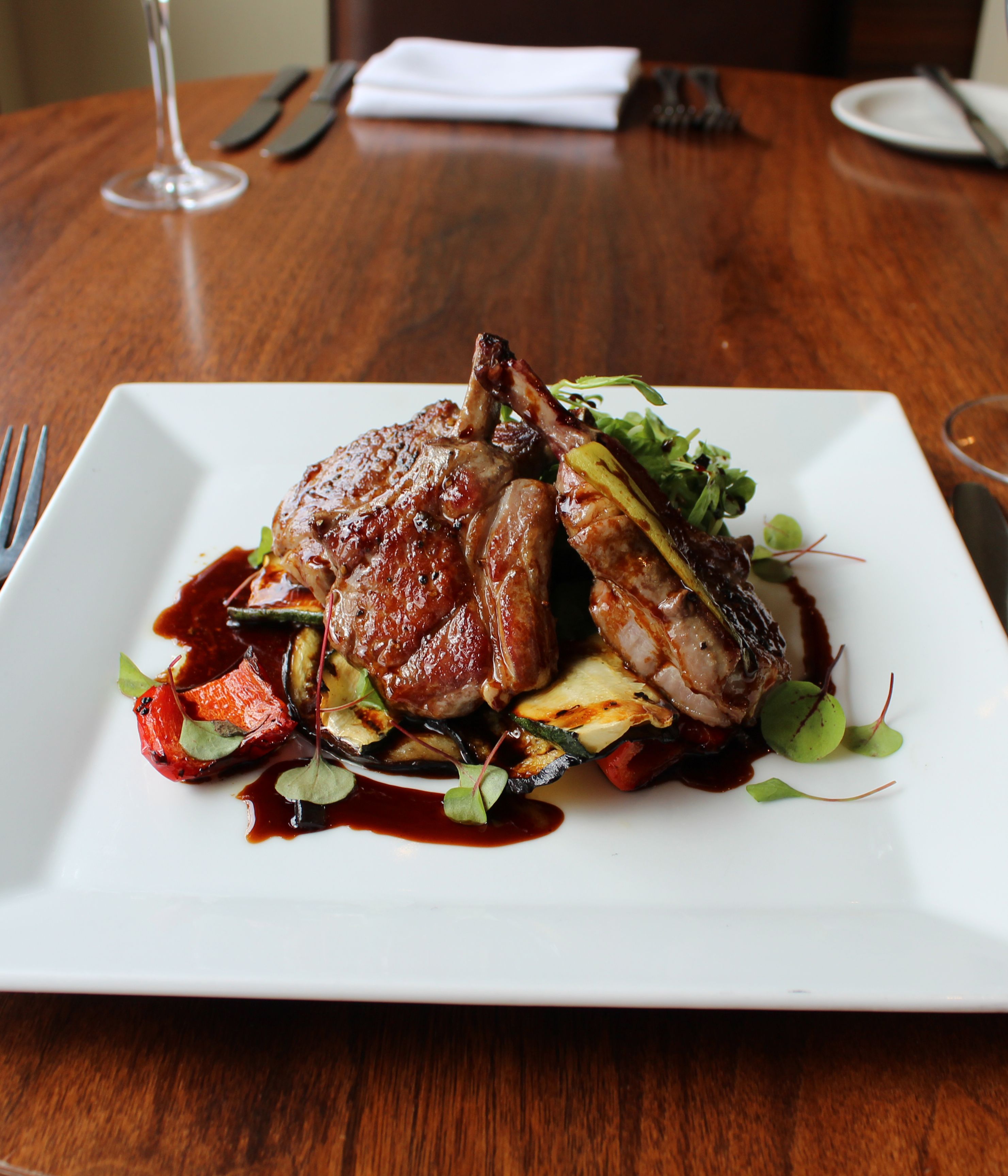 Gourmet lamb chops served on a white square plate with roasted vegetables and sauce, on a wooden restaurant table.