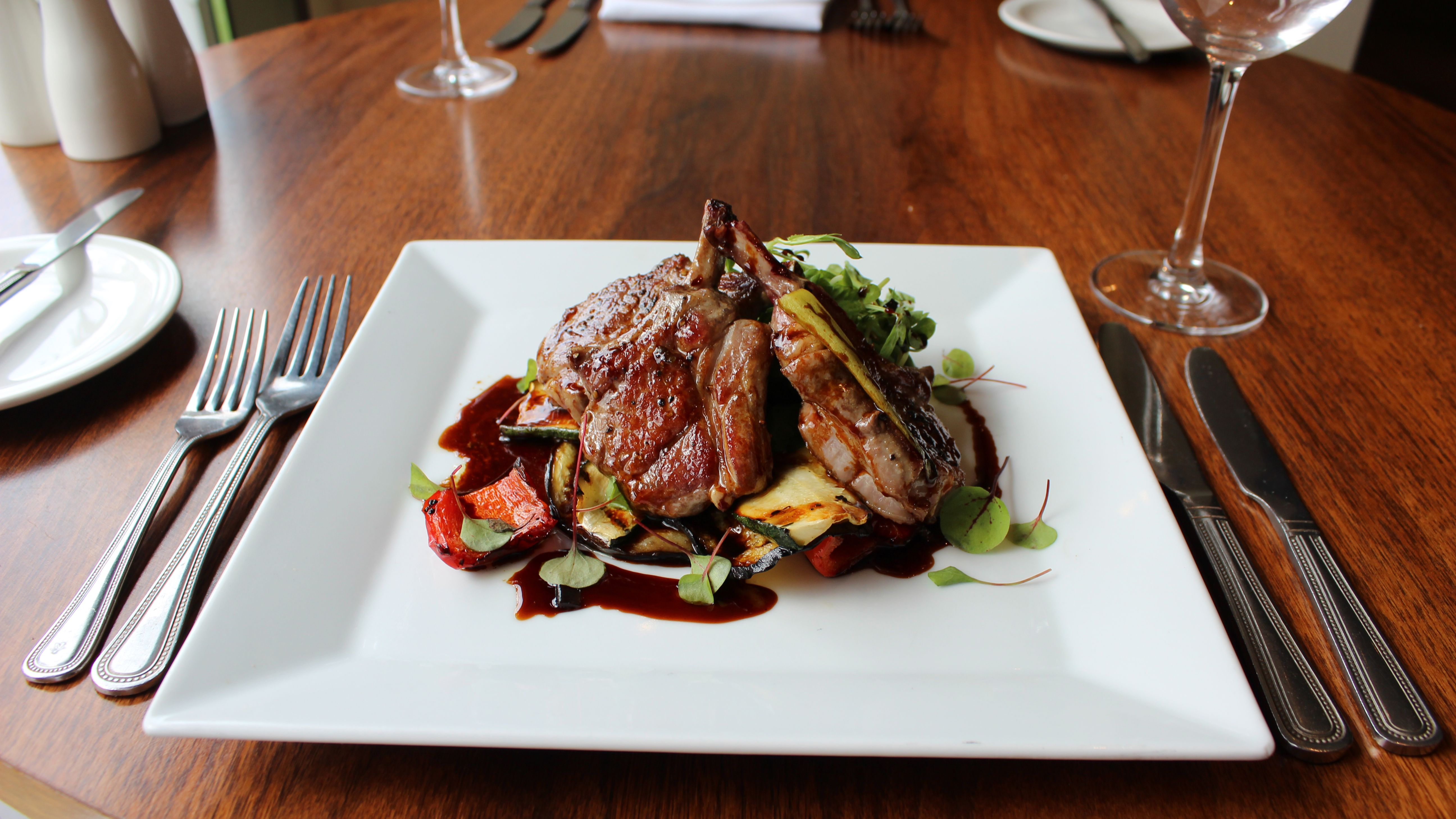 Gourmet lamb chops served on a white square plate with roasted vegetables and sauce, on a wooden restaurant table.