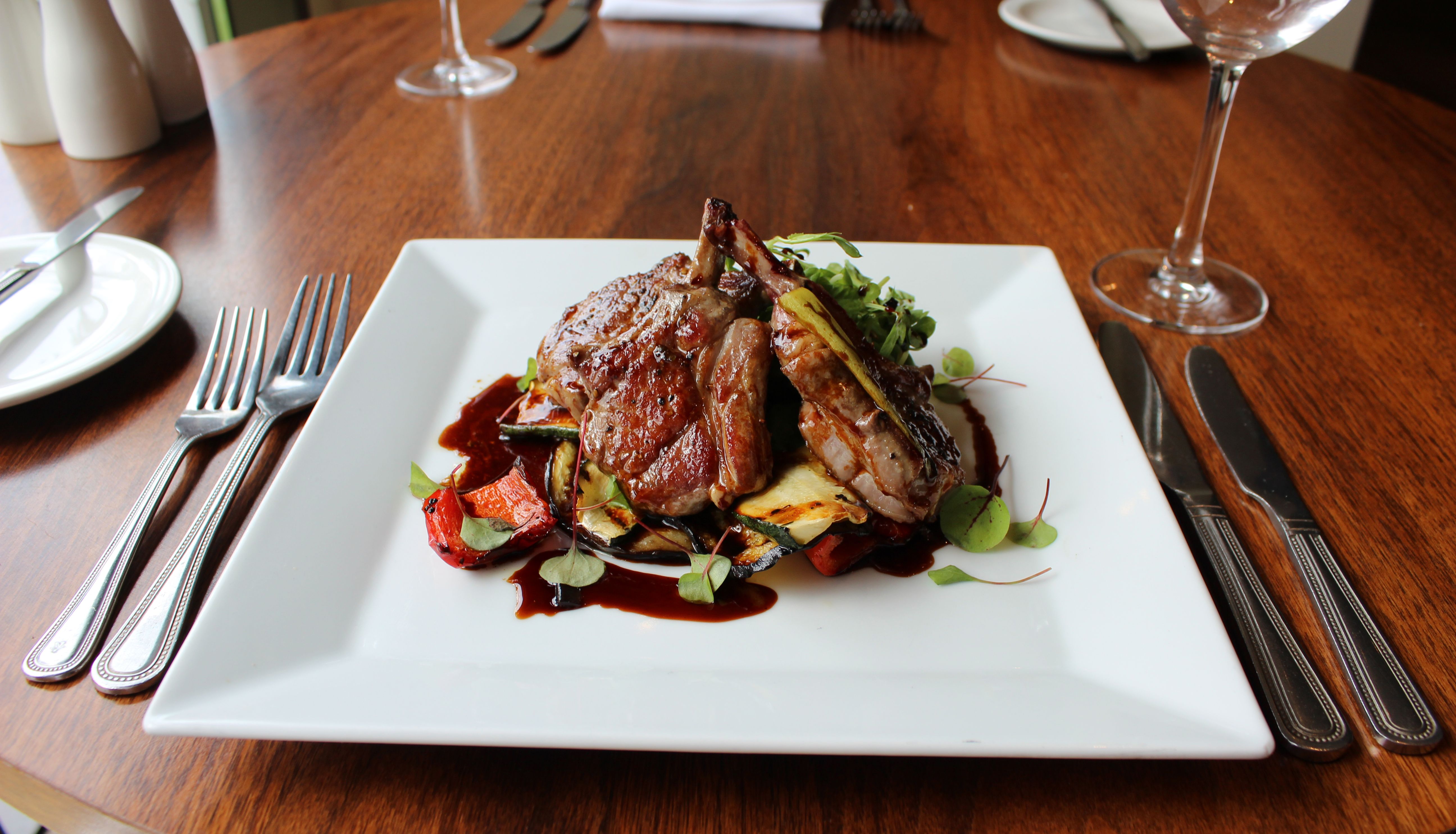 Gourmet lamb chops served on a white square plate with roasted vegetables and sauce, on a wooden restaurant table.
