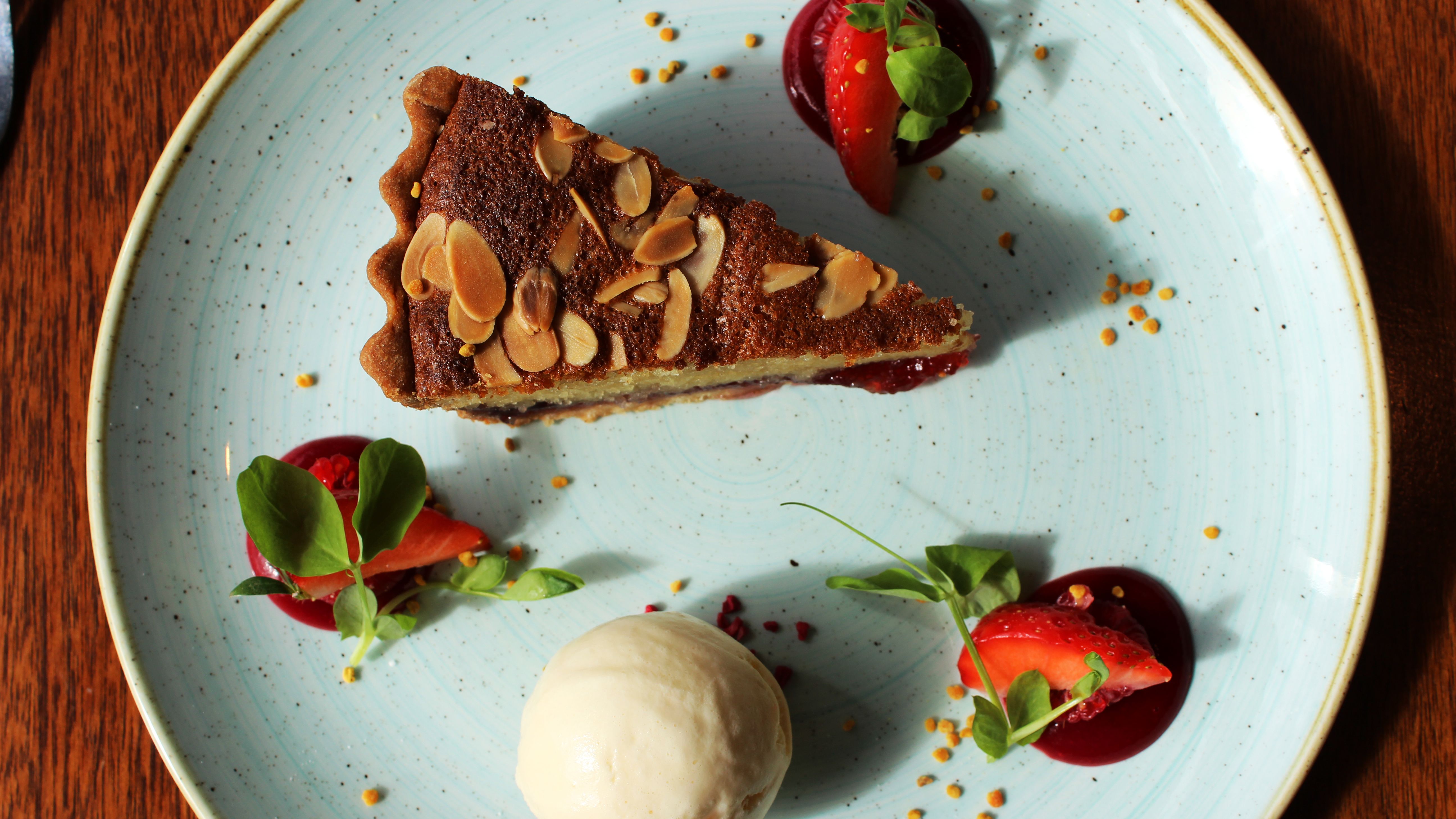 A slice of almond cake served with vanilla ice cream, strawberry slices, berry sauce, and microgreens on a light blue plate.