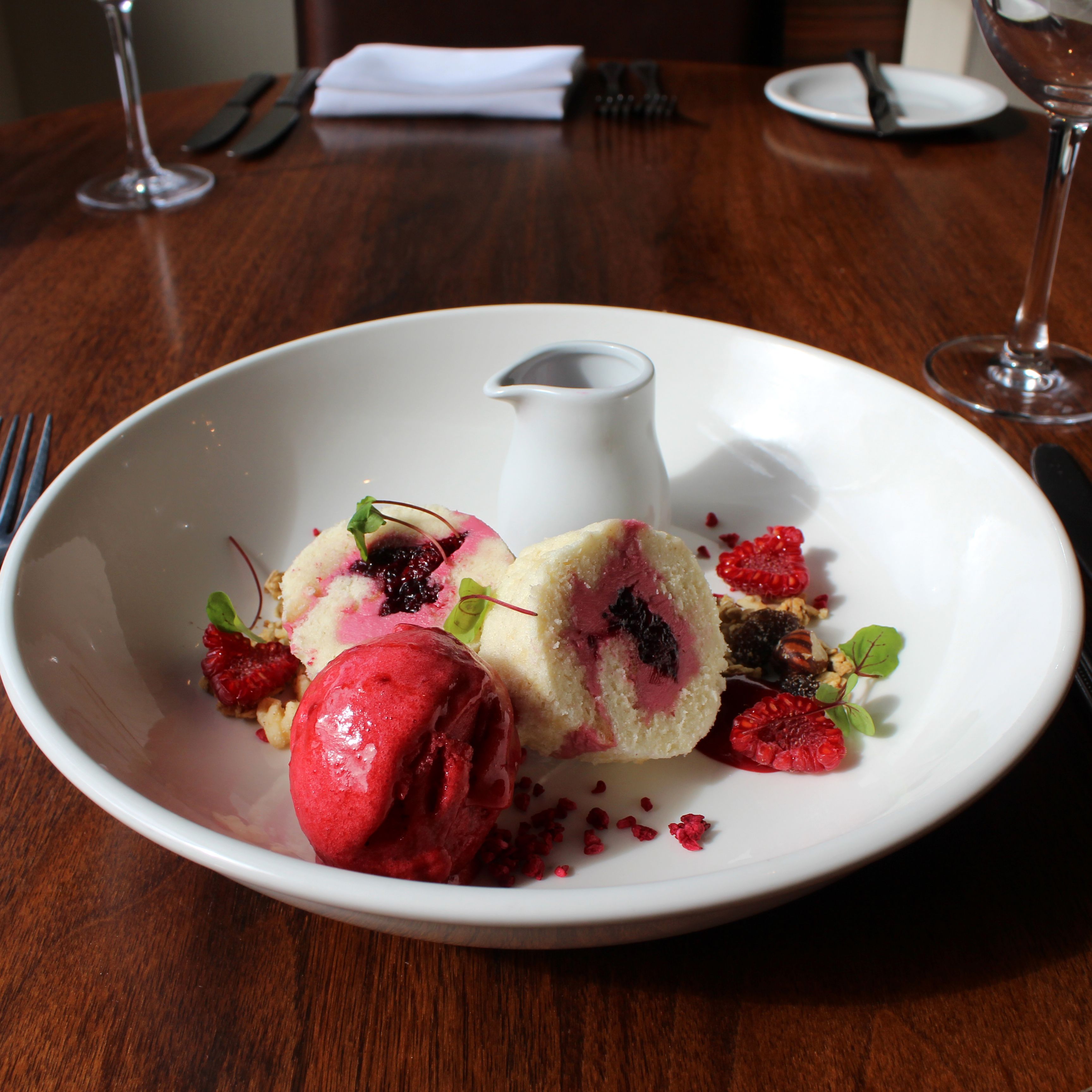 Plated dessert featuring scoops of berry sorbet, pieces of a rolled sponge cake with berry filling, garnished with nuts and fresh raspberries on a restaurant table.