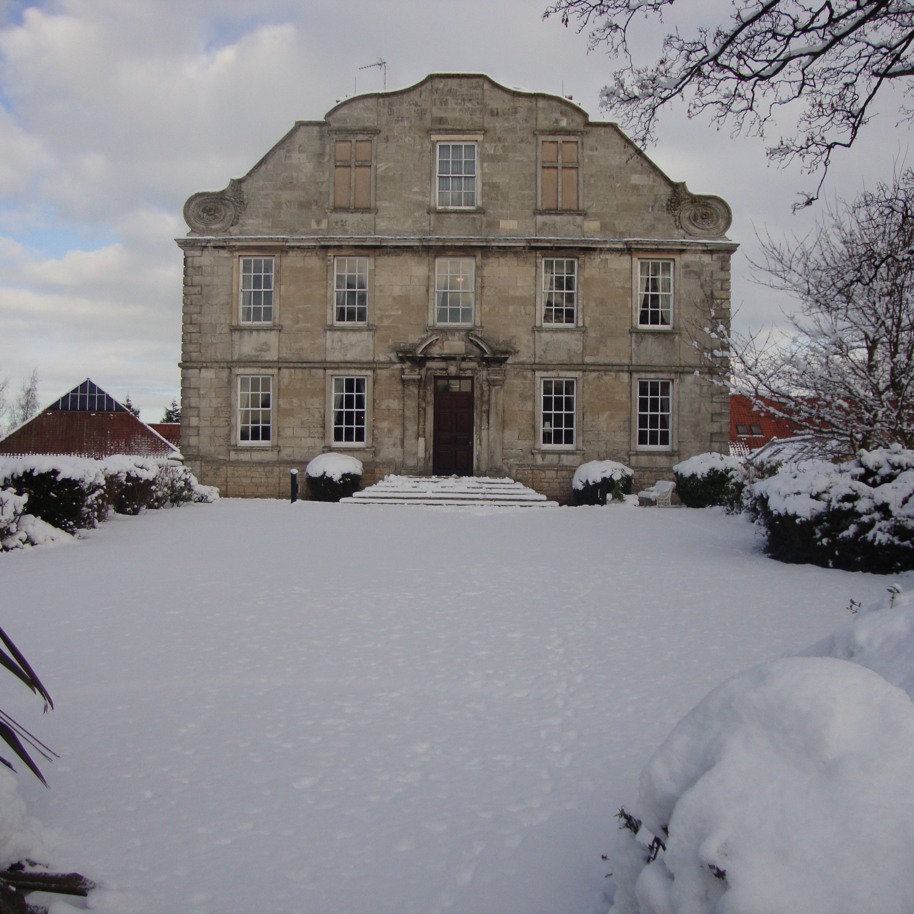 Historic stone building with multiple windows, surrounded by a snowy landscape and bushes covered in snow.