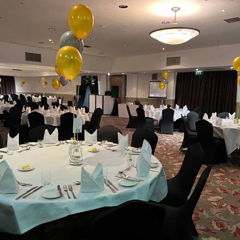 Elegant banquet hall with round tables covered with white tablecloths, black chairs, and gold and silver balloons.