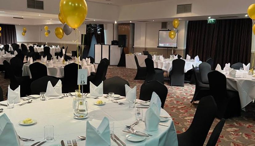 Elegant banquet hall with round tables covered with white tablecloths, black chairs, and gold and silver balloons.