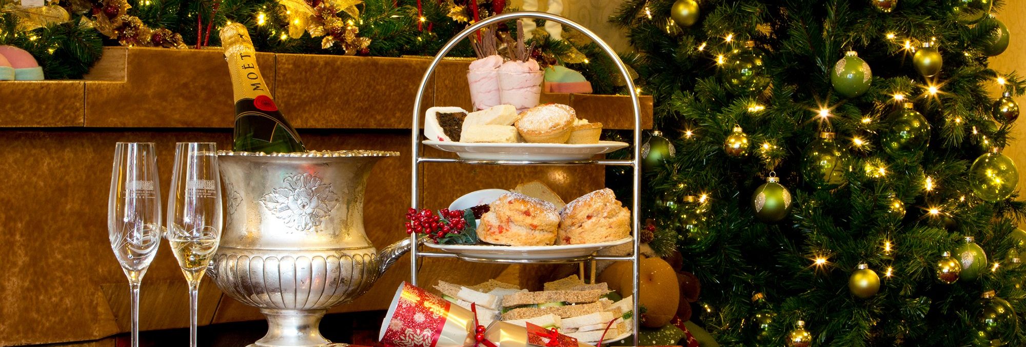 Festive Christmas tea setup with pastries, champagne, and decorations by a Christmas tree.