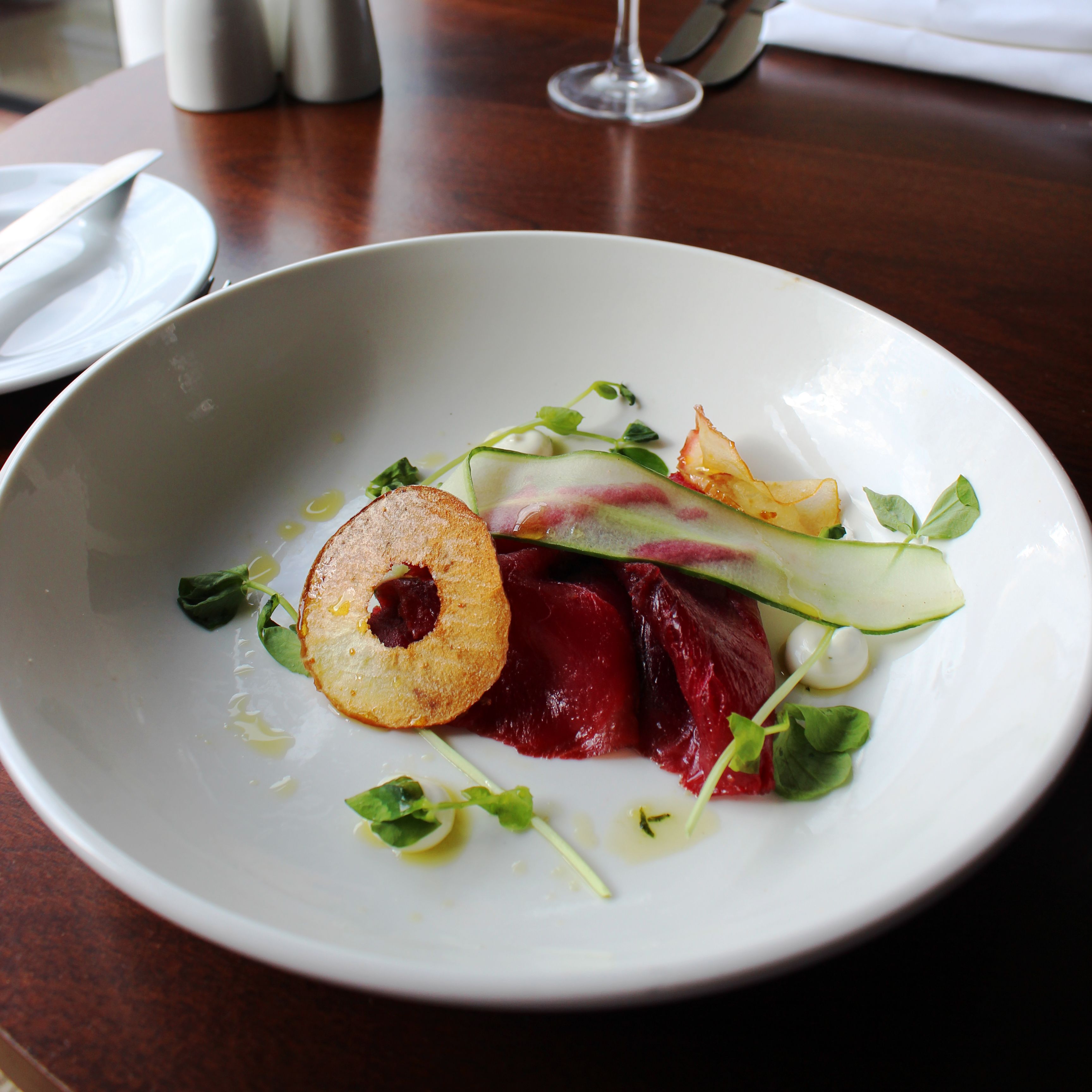 Elegant plated dish with thinly sliced vegetables, a crisp potato round, and greens in a white bowl on a restaurant table.