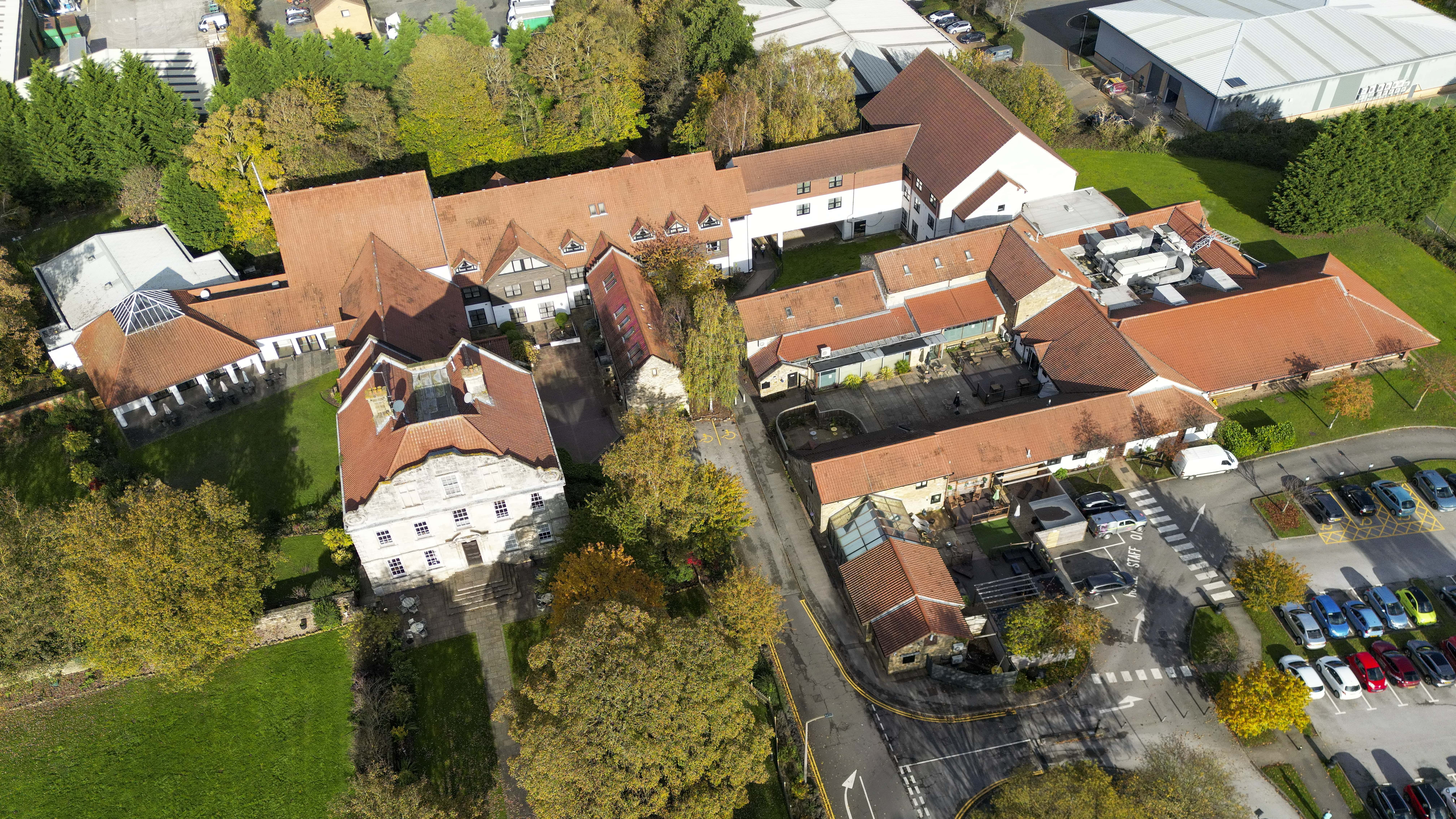 Aerial view of a large building complex with red-tiled roofs surrounded by greenery and a parking lot.