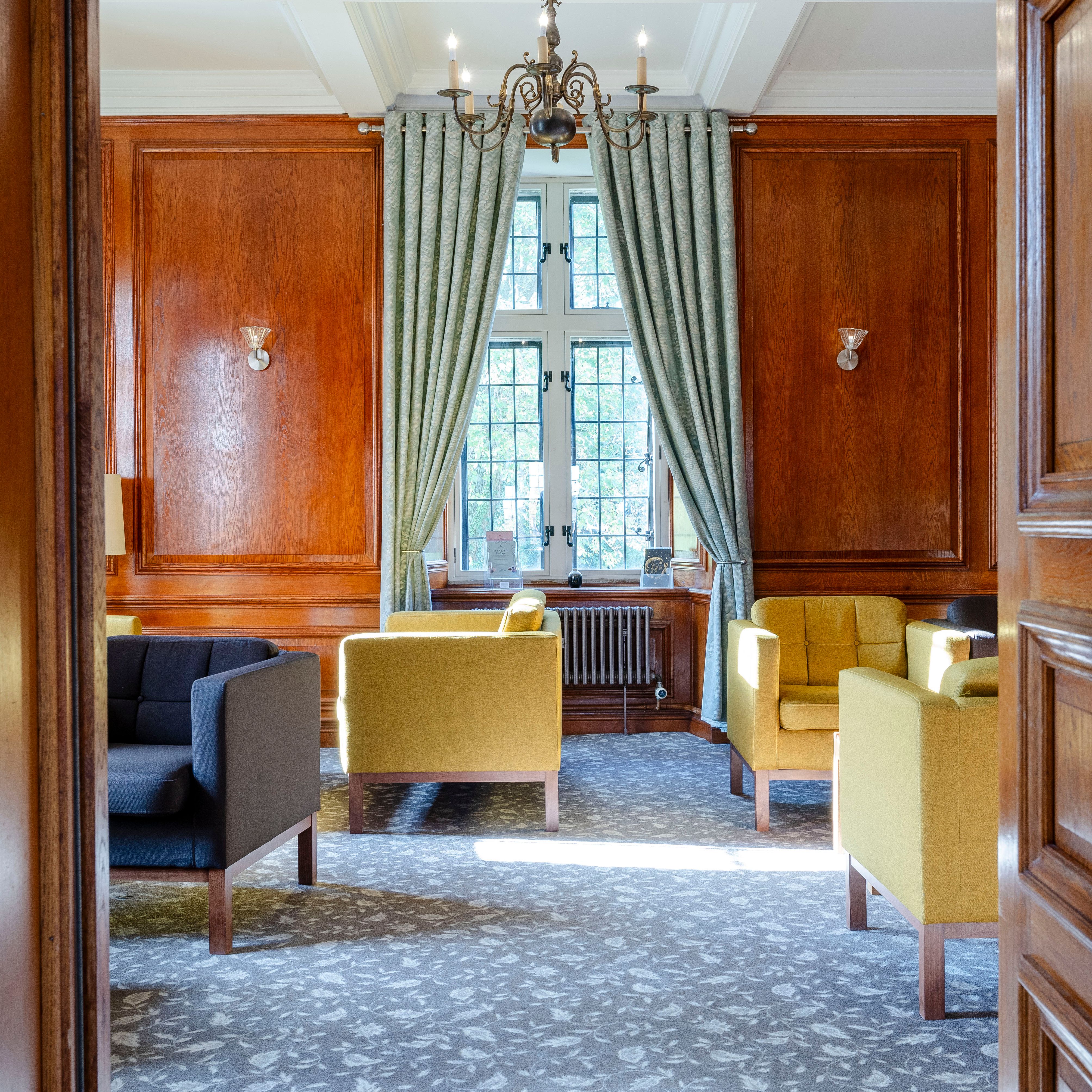 Elegant lounge with yellow and grey armchairs, wooden paneled walls, a large window with curtains, and a chandelier.