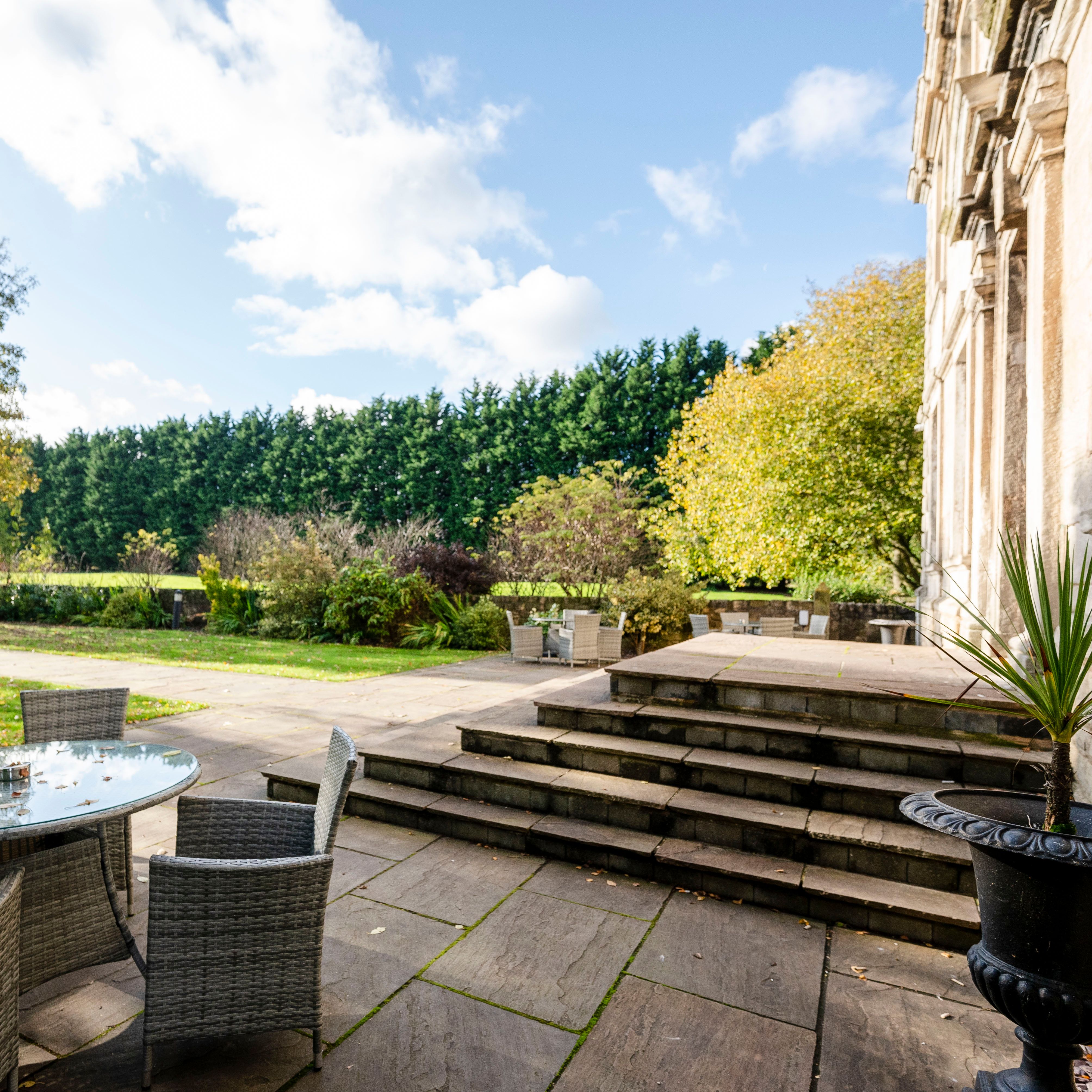Outdoor patio area with wicker tables and chairs, surrounded by greenery and trees on a sunny day