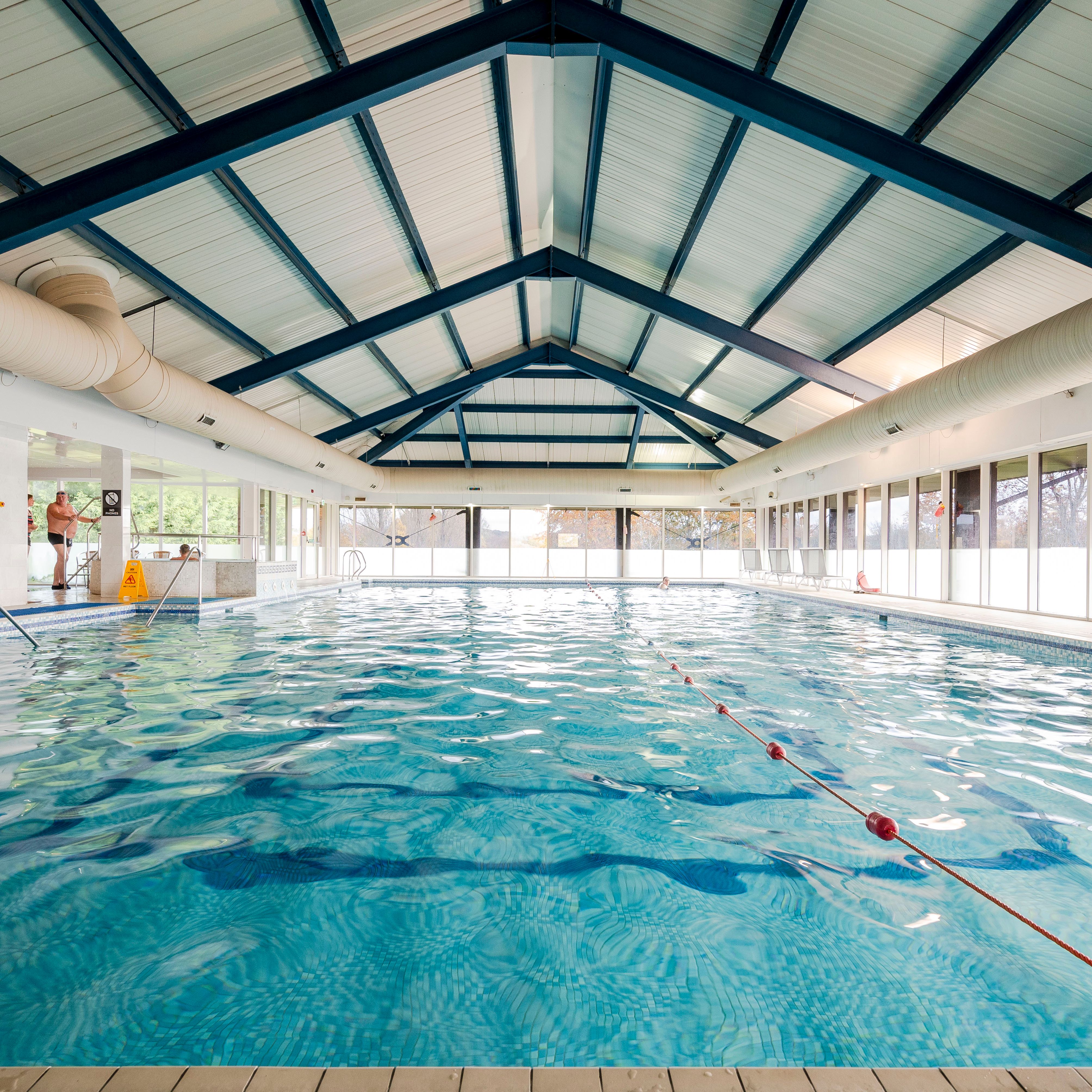 Indoor swimming pool with blue water, a high ceiling, and large windows along the sides.