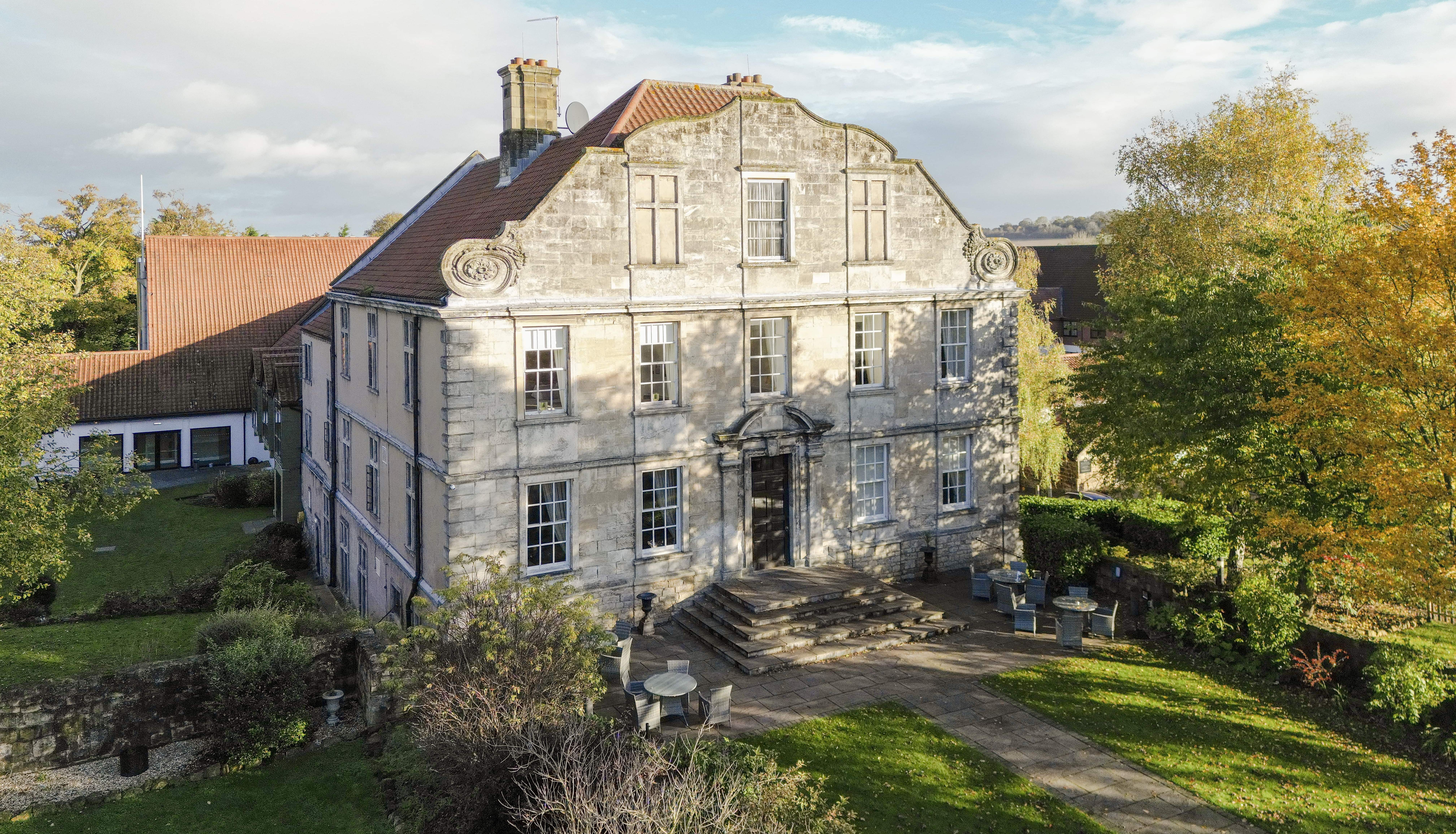 Aerial view of an old stone mansion with a grand staircase and red tiled roof surrounded by gardens and trees.