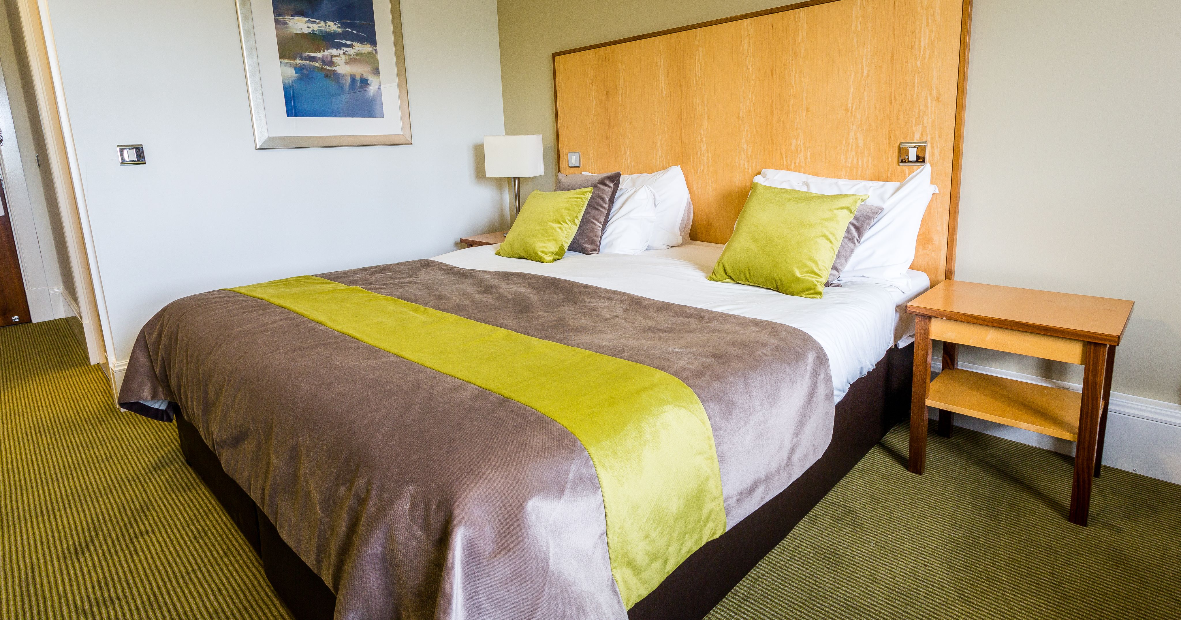 A neatly made bed with green accent pillows and a green and brown bed runner in a well-lit hotel room.