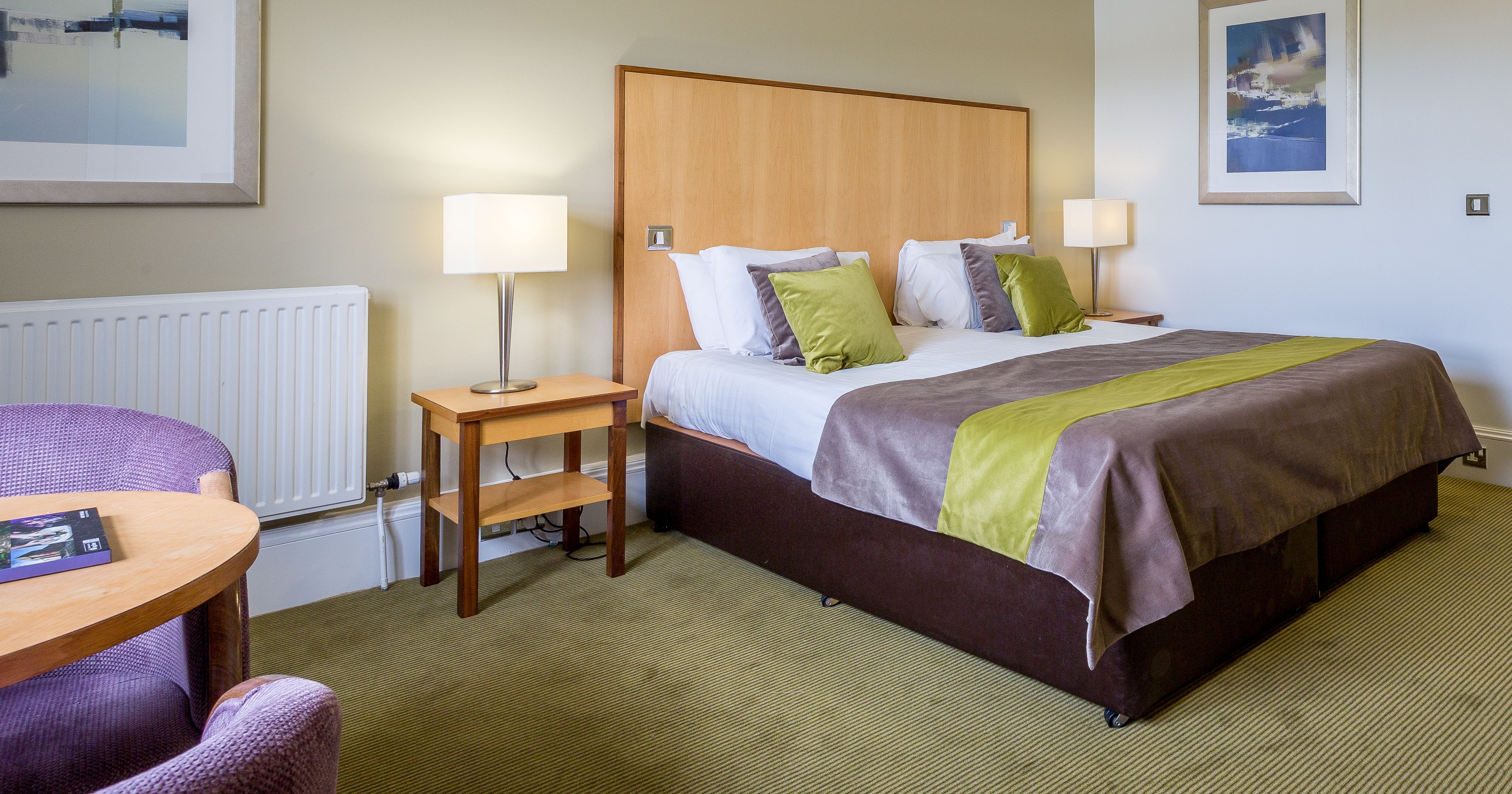 Modern hotel room with a large bed, green and gray decor, bedside tables with lamps, and framed artwork on the walls.