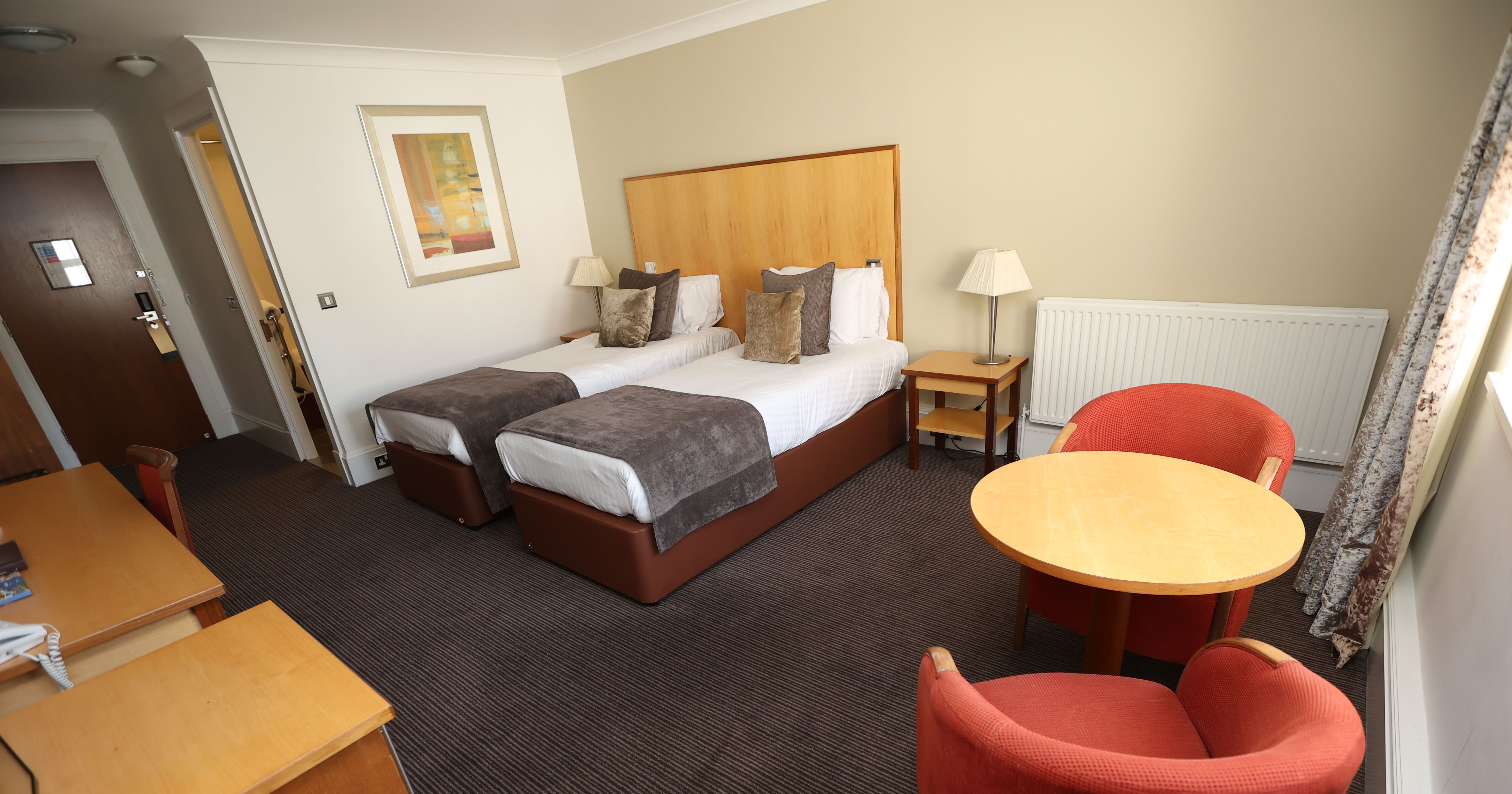 Hotel room with two single beds, a table with two red chairs, and a desk.