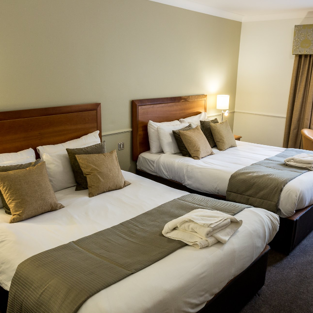 A hotel room with two queen-sized beds, each with multiple pillows, bedside tables with lamps, a window with curtains, and a small seating area.