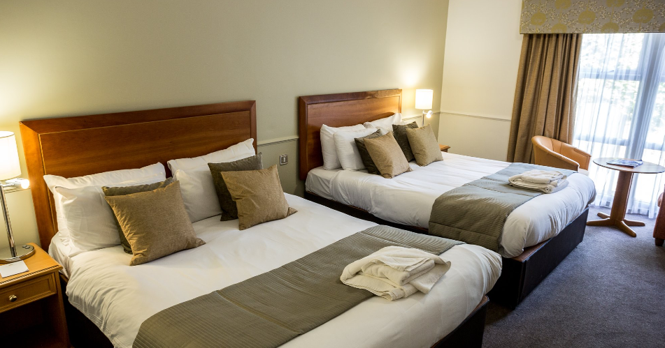 A hotel room with two queen-sized beds, each with multiple pillows, bedside tables with lamps, a window with curtains, and a small seating area.