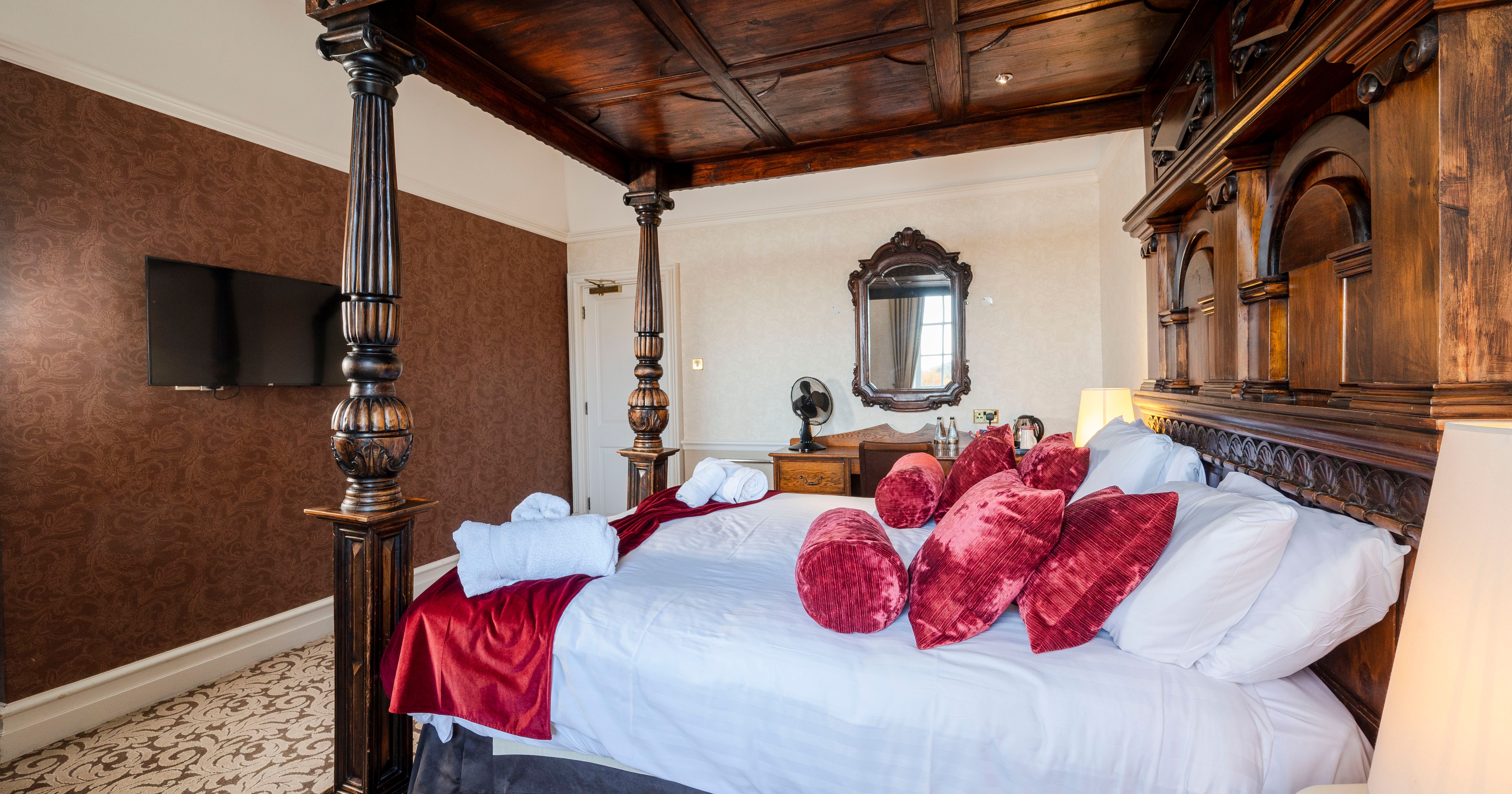 Elegant hotel room with a large carved wood four-poster bed, white linens, red velvet pillows, and a vintage vanity with mirror.