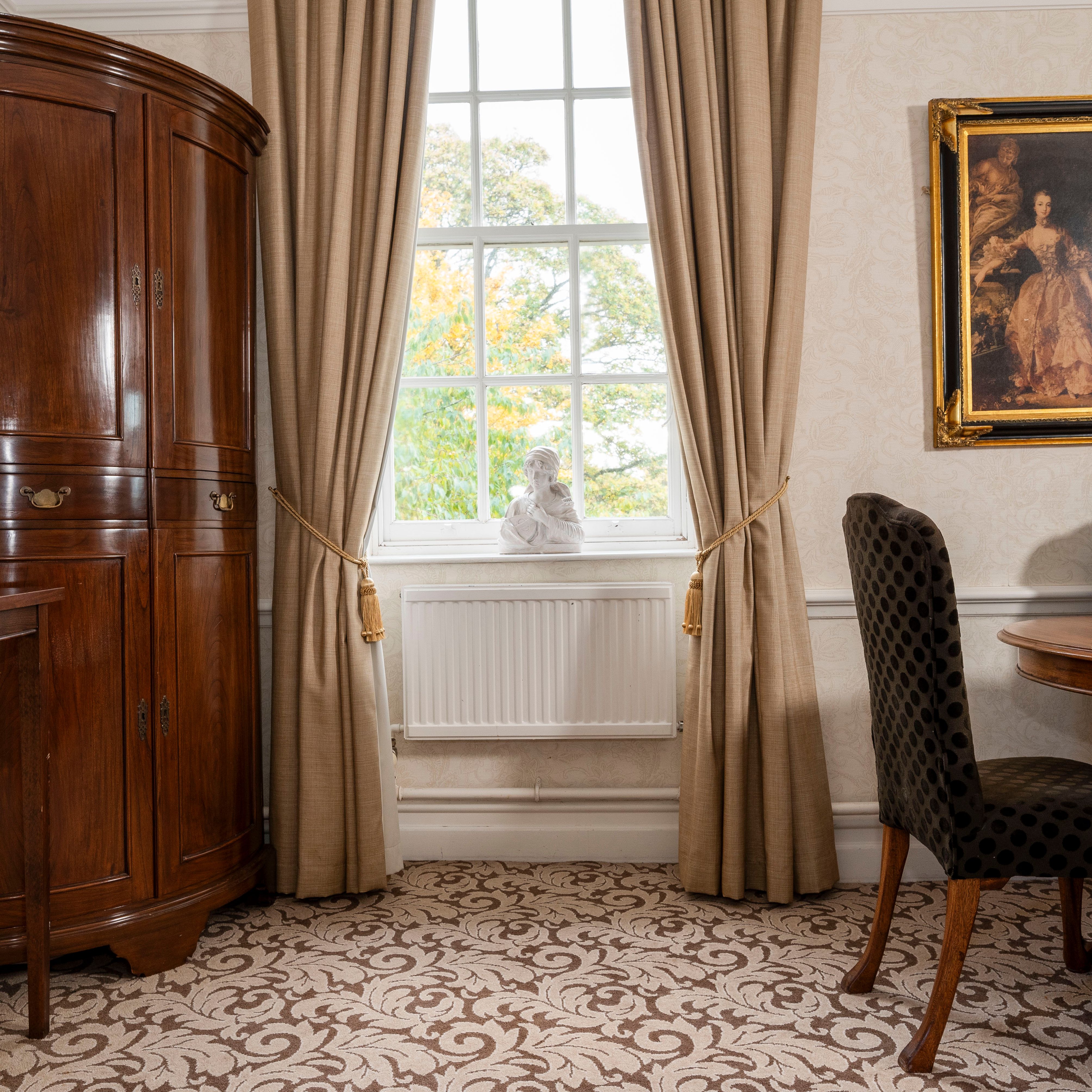 Elegant traditional room with wooden furniture, a window with beige curtains, framed artwork, and a bust on the windowsill.