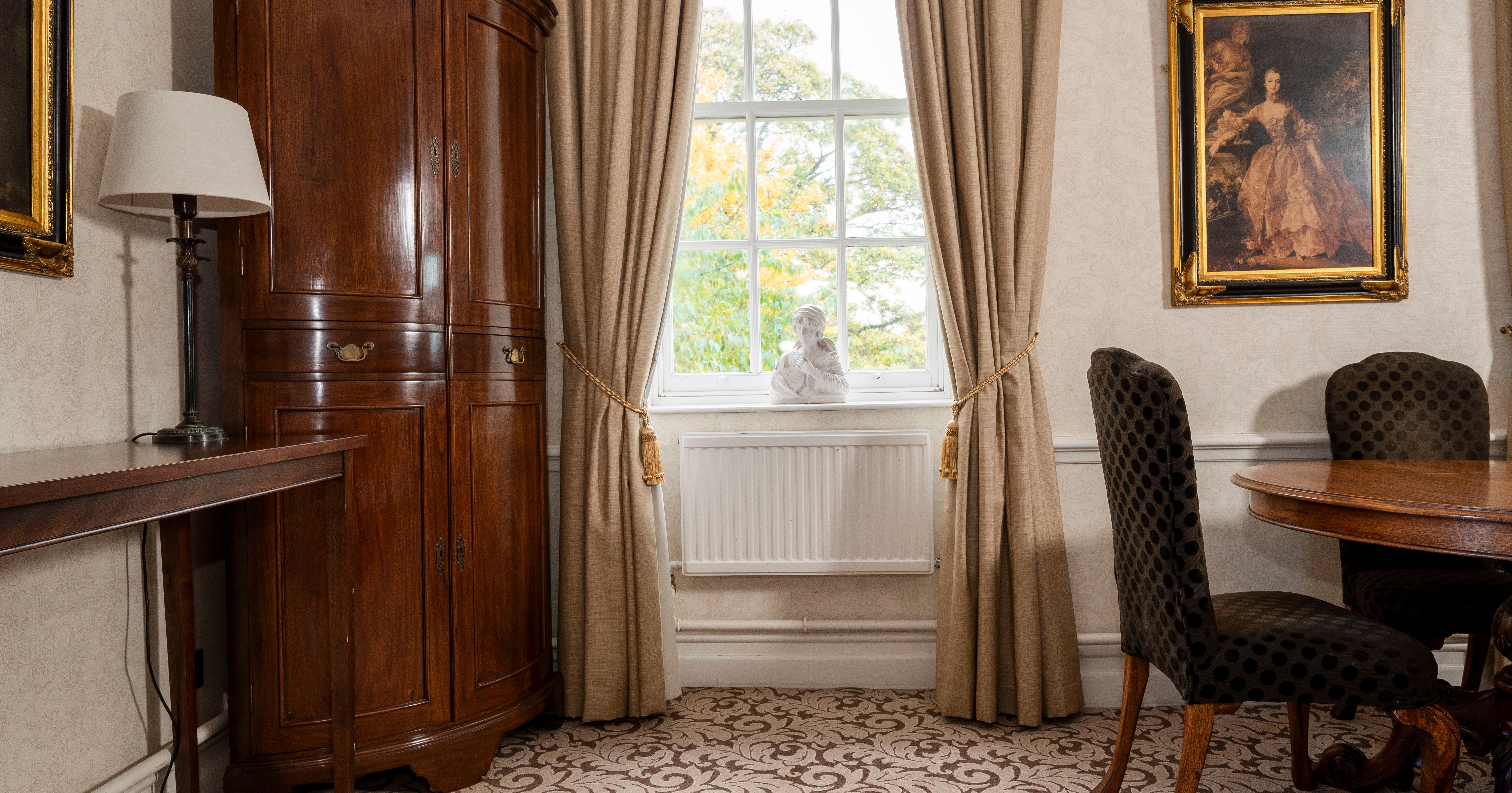 Elegant traditional room with wooden furniture, a window with beige curtains, framed artwork, and a bust on the windowsill.