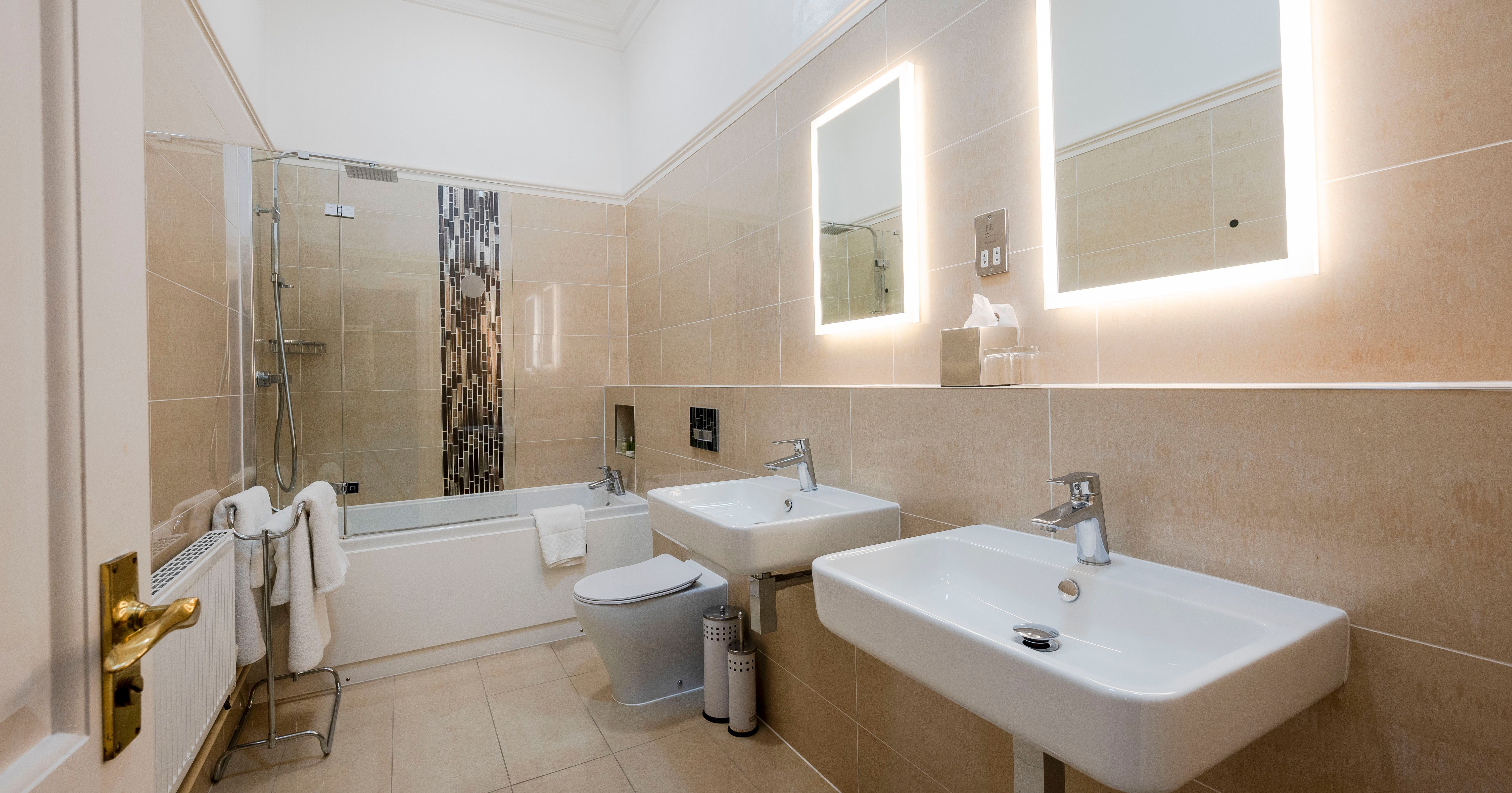 Modern bathroom with two sinks, illuminated mirrors, and a bathtub with glass shower screen