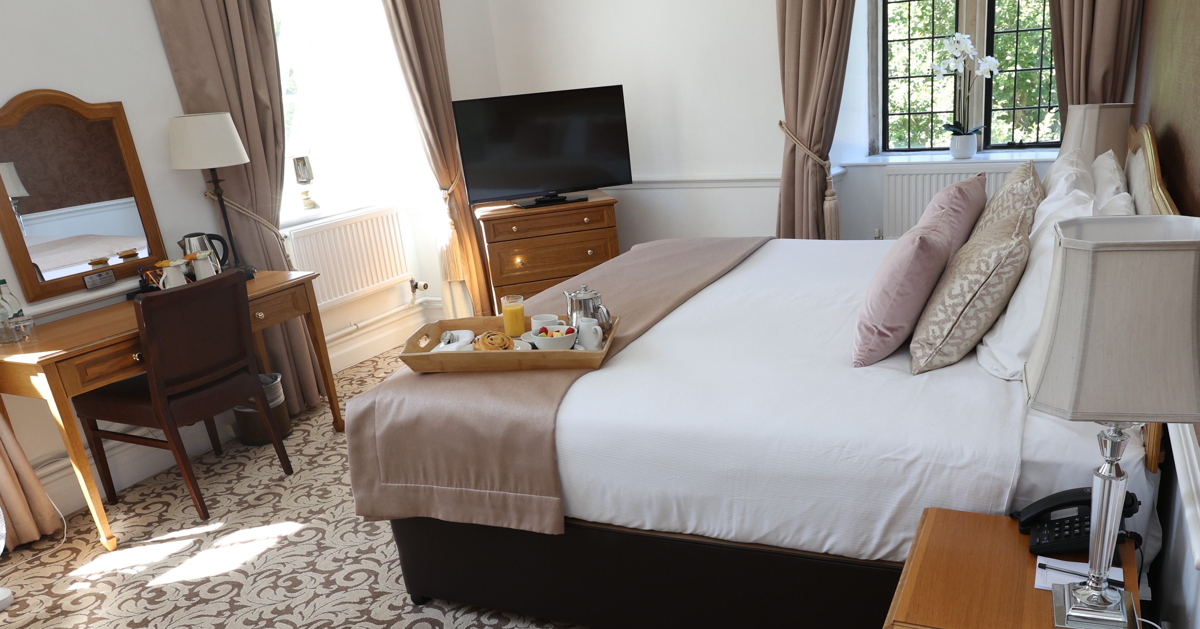Elegant hotel room with a neatly made bed, breakfast tray, TV, desk, and large windows with curtains.