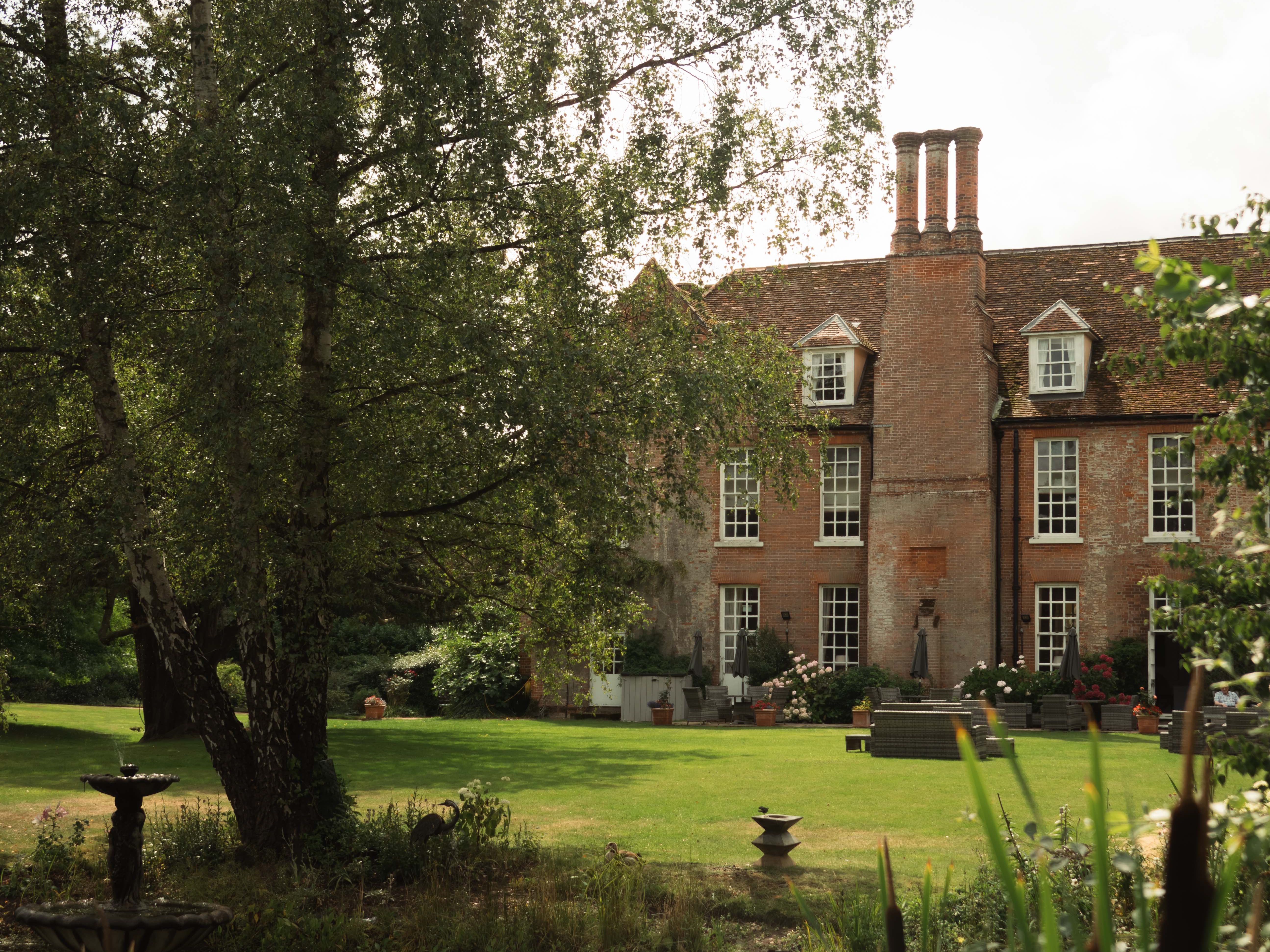 Brick English manor house with tall windows and chimneys, seen across a pond and lawn with a fountain and trees.