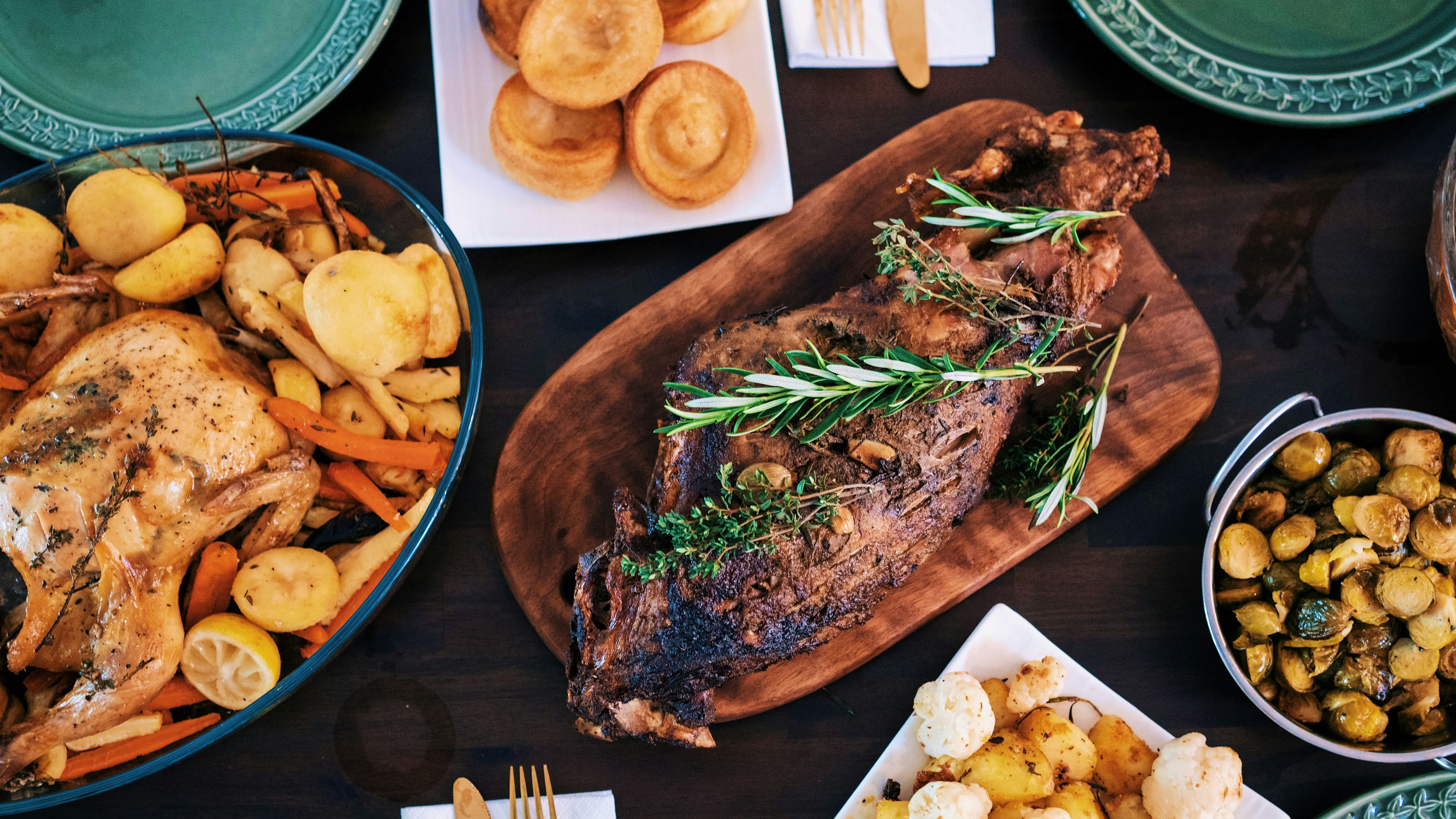 A table set with a roasted chicken, a seasoned roast with herbs, Yorkshire puddings, brussels sprouts, cauliflower and potatoes, and green plates