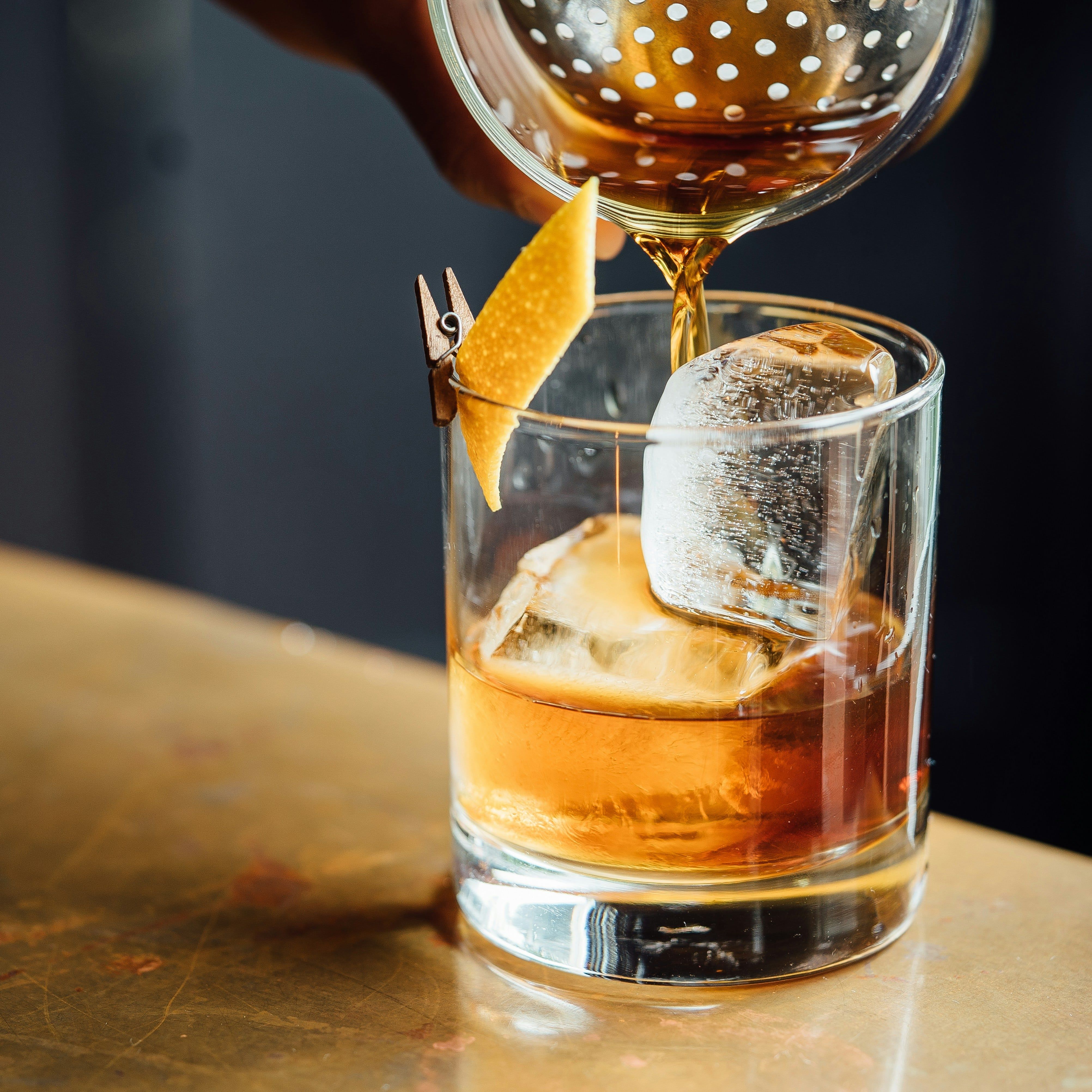 Whiskey cocktail being poured into a glass with ice and an orange peel garnish.