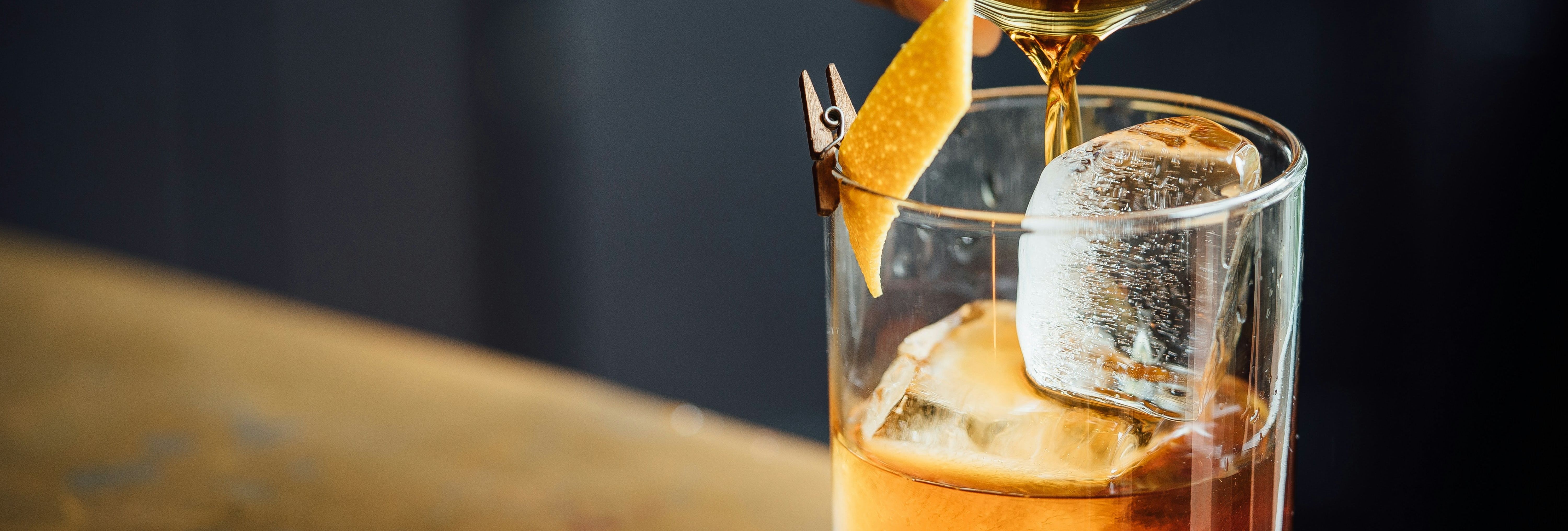 Whiskey cocktail being poured into a glass with ice and an orange peel garnish.