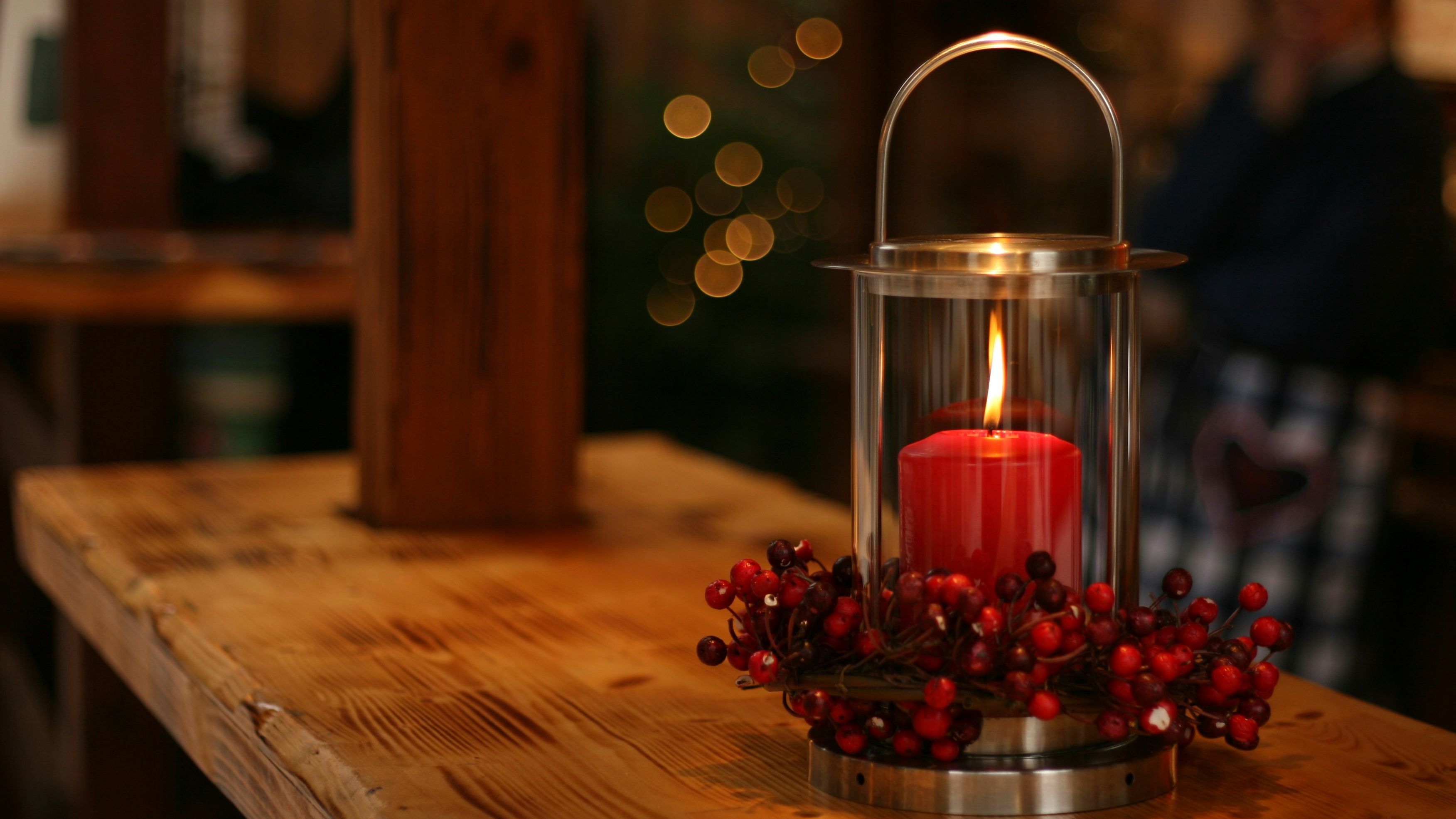 Red candle burning inside a glass lantern decorated with red berries, placed on a wooden table.