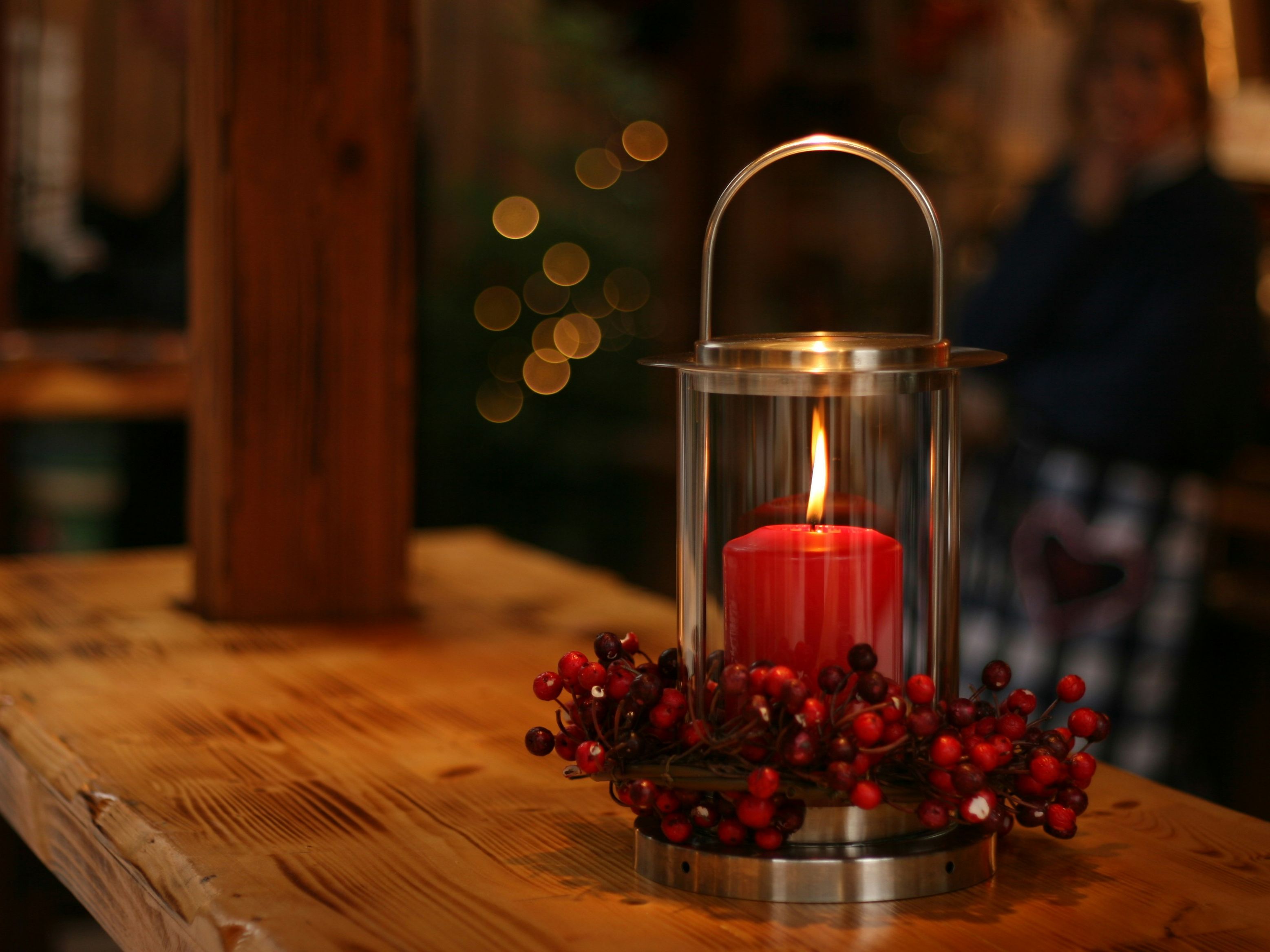 Red candle burning inside a glass lantern decorated with red berries, placed on a wooden table.