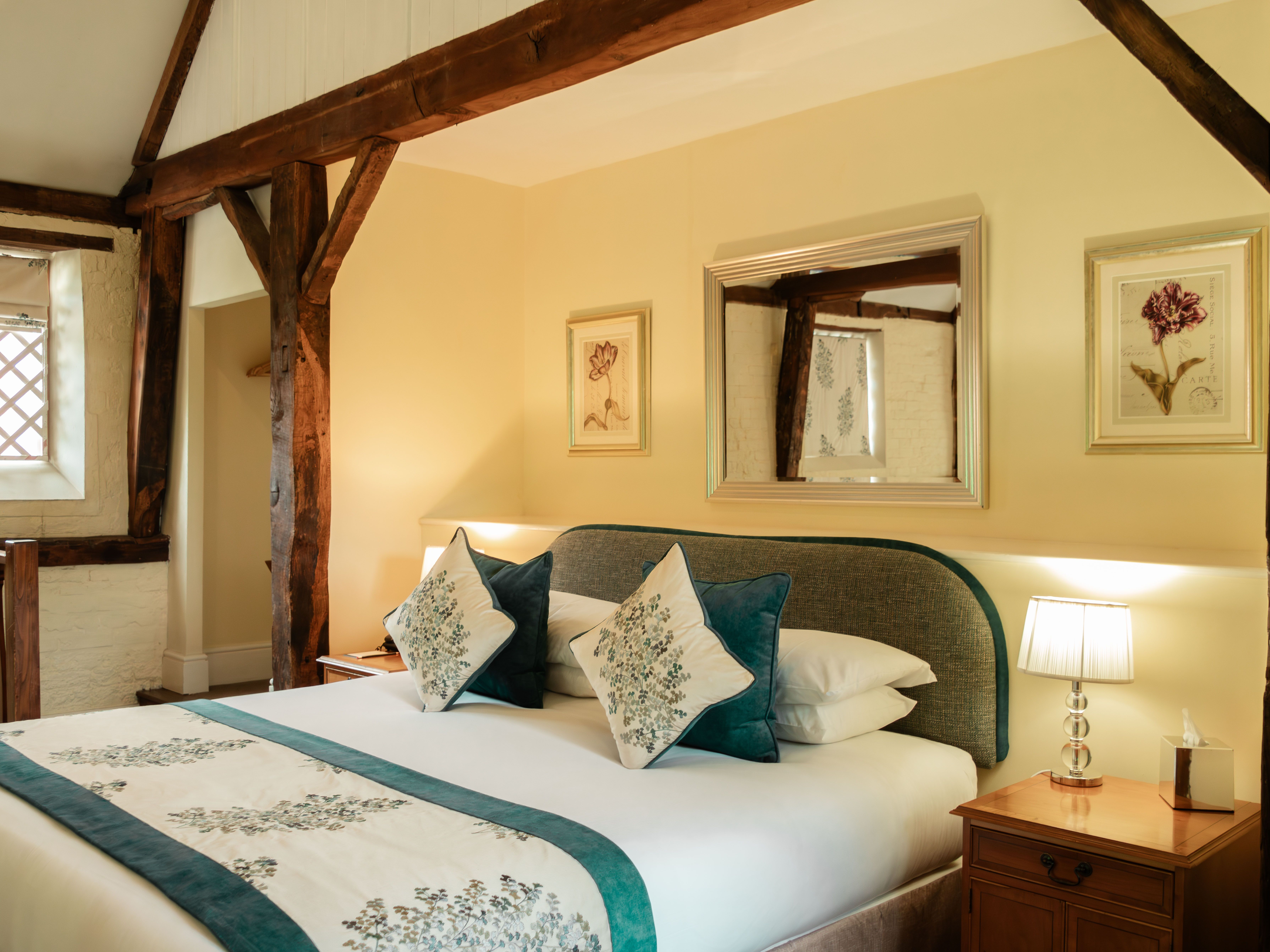 Cozy bedroom with wooden beams, a neatly made bed, bedside table, and decorative artwork on the wall.