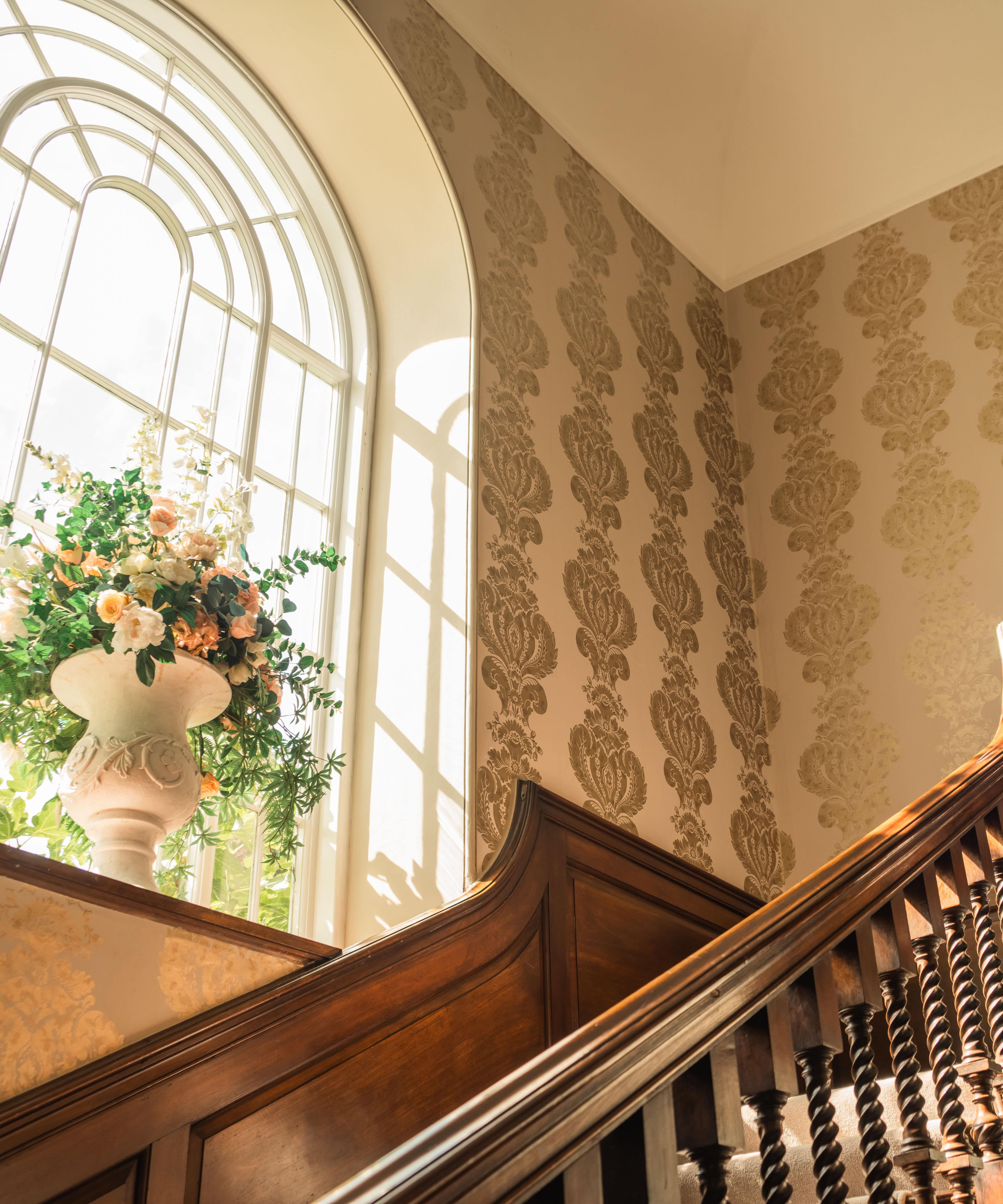 Elegant wooden staircase with ornate railing and a large flower arrangement by a tall arched window