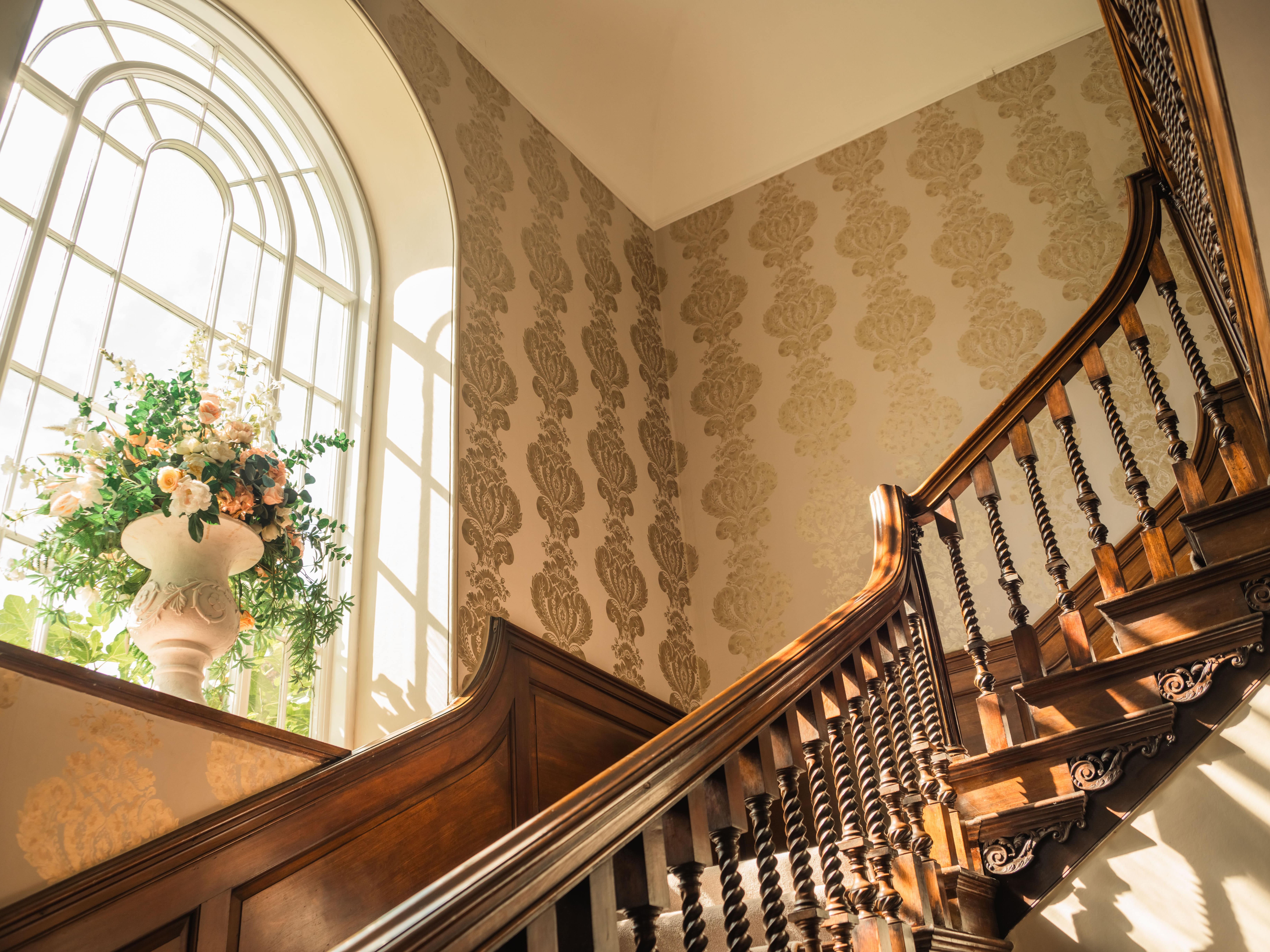 Elegant wooden staircase with ornate railing beside a large arched window and a floral arrangement in a white vase.