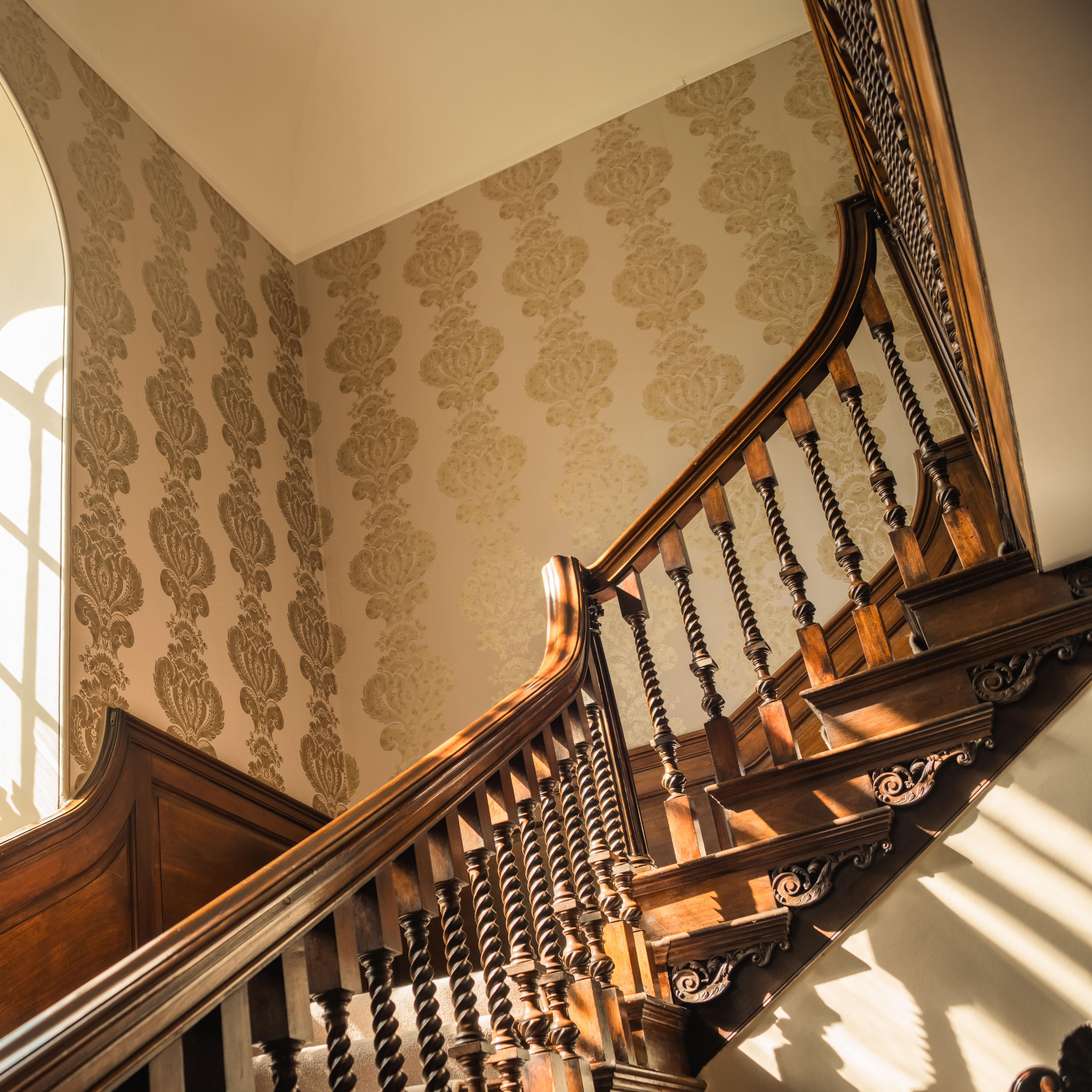 Elegant wooden staircase with ornate railing beside a large arched window and a floral arrangement in a white vase.
