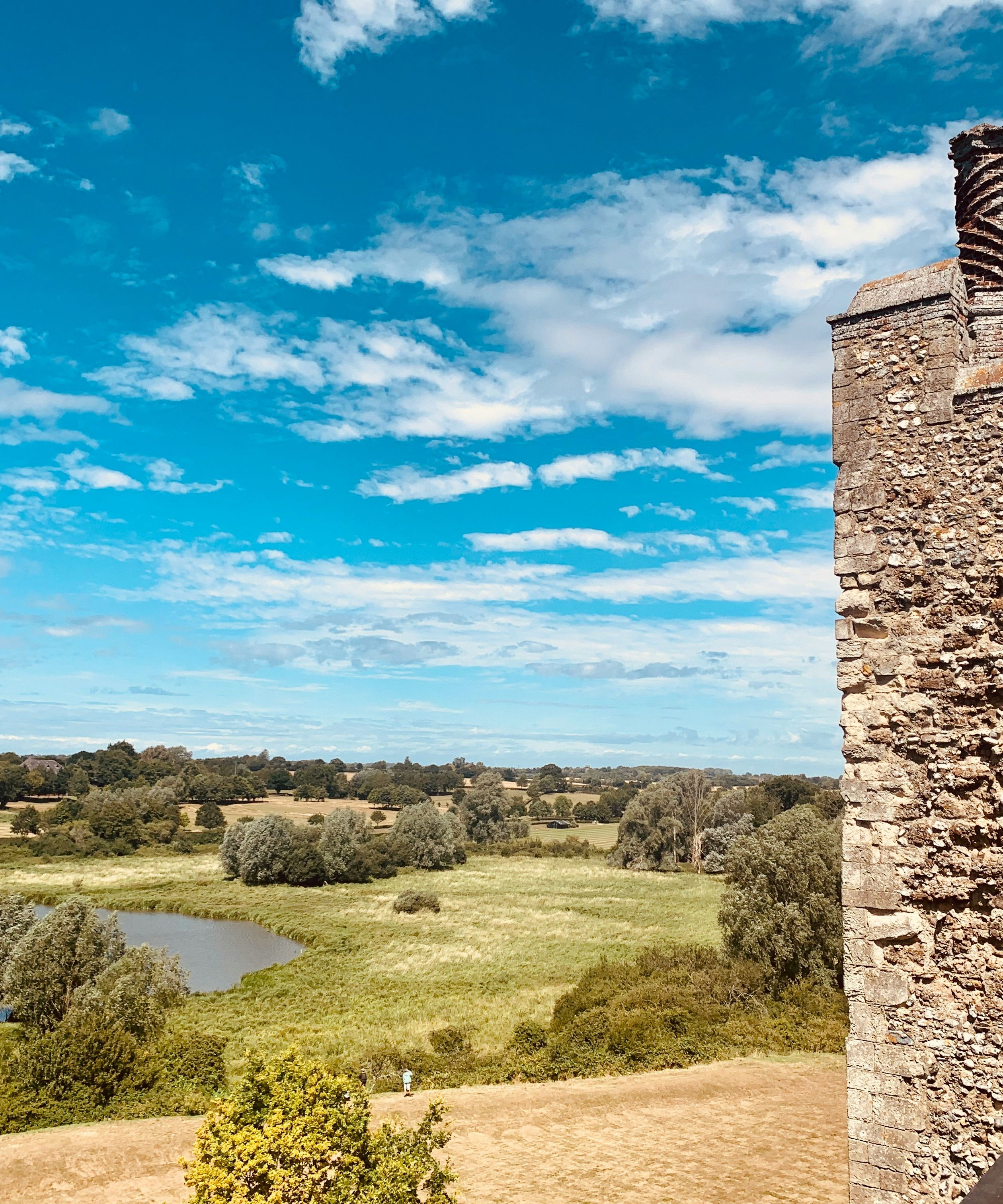Stone castle wall overlooking a green landscape with a pond and trees under a blue sky with clouds.