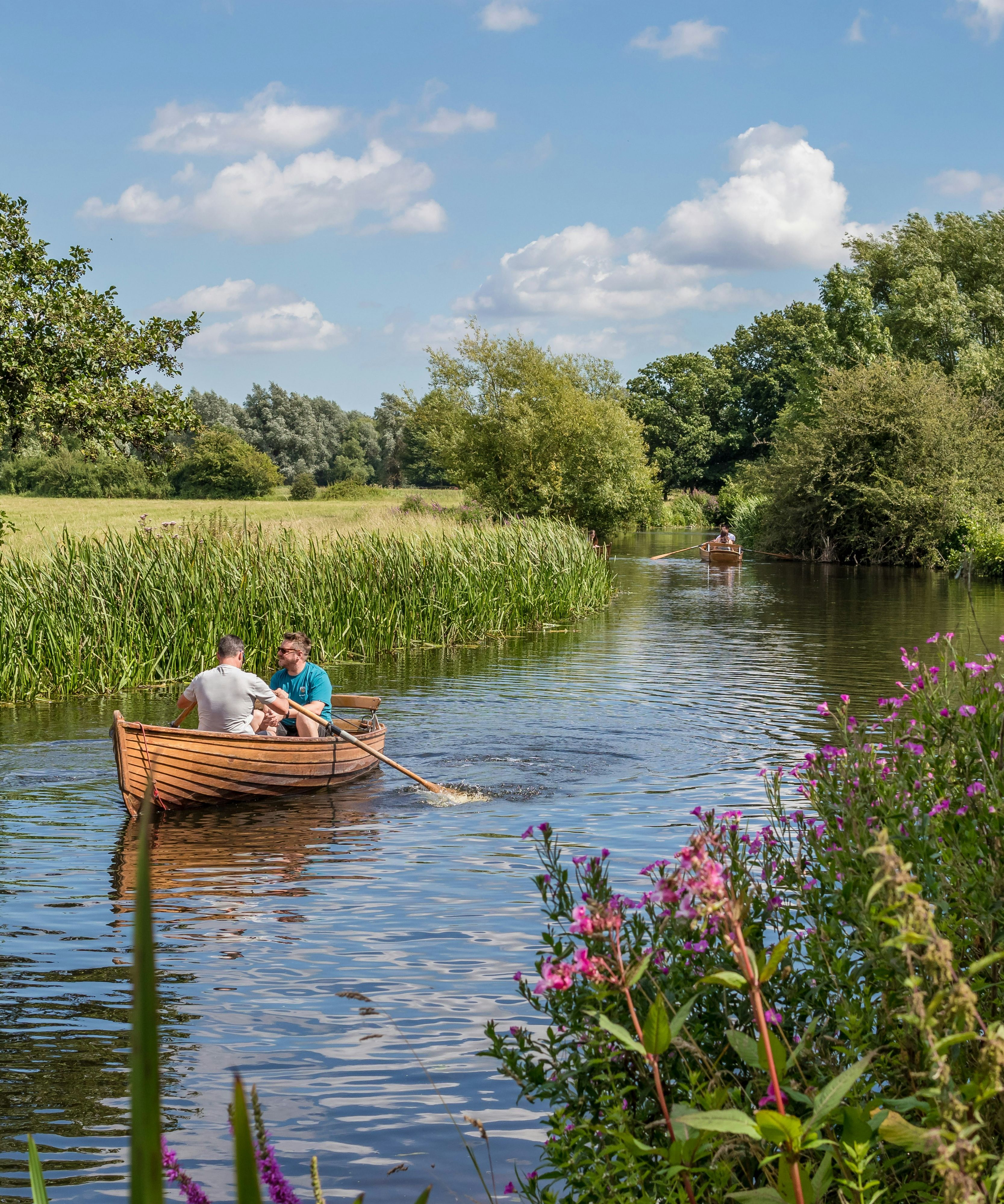 Two people rowing a wooden boat on a calm river surrounded by greenery and wildflowers.