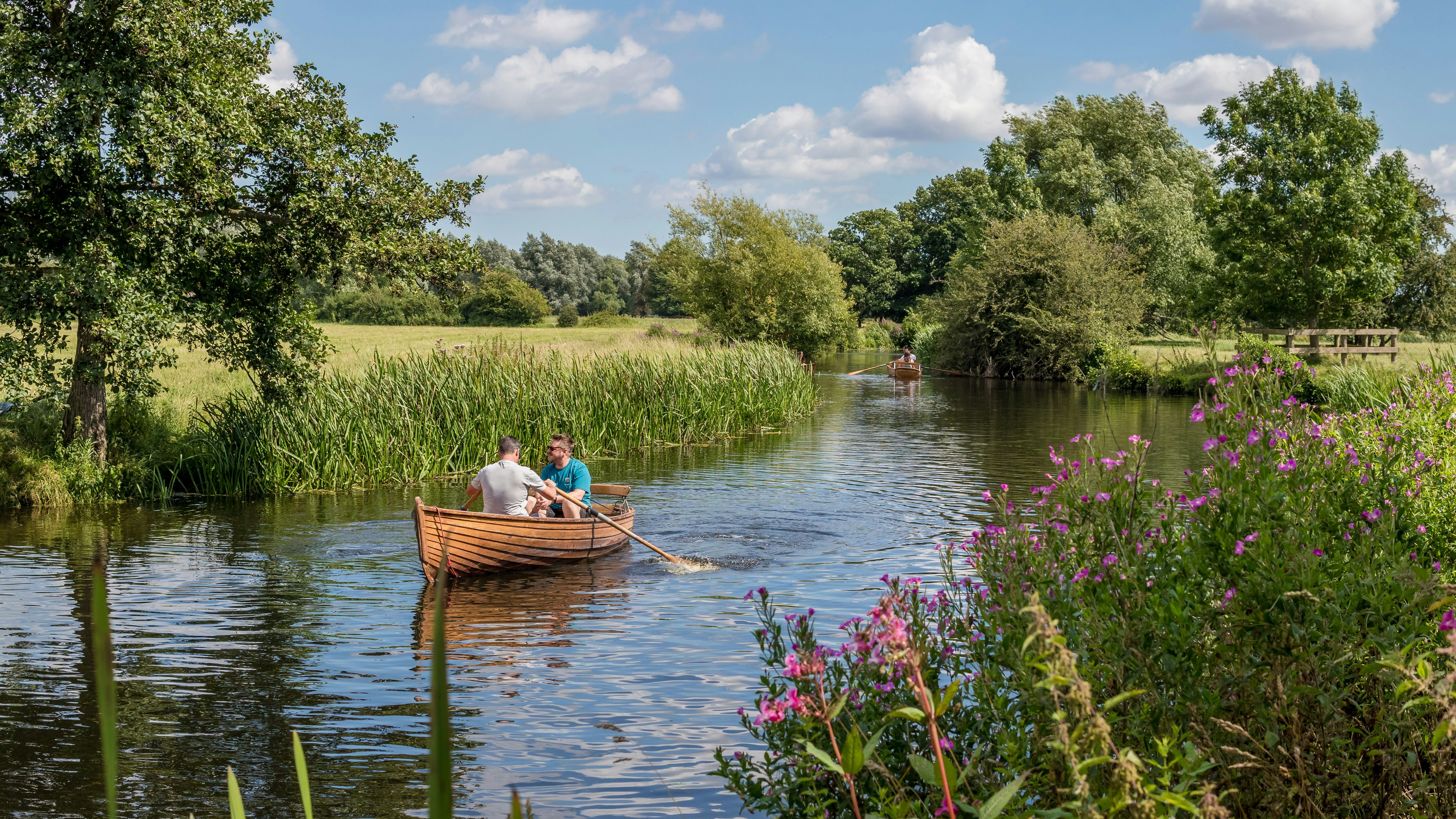 Two people rowing a wooden boat on a calm river surrounded by greenery and wildflowers.