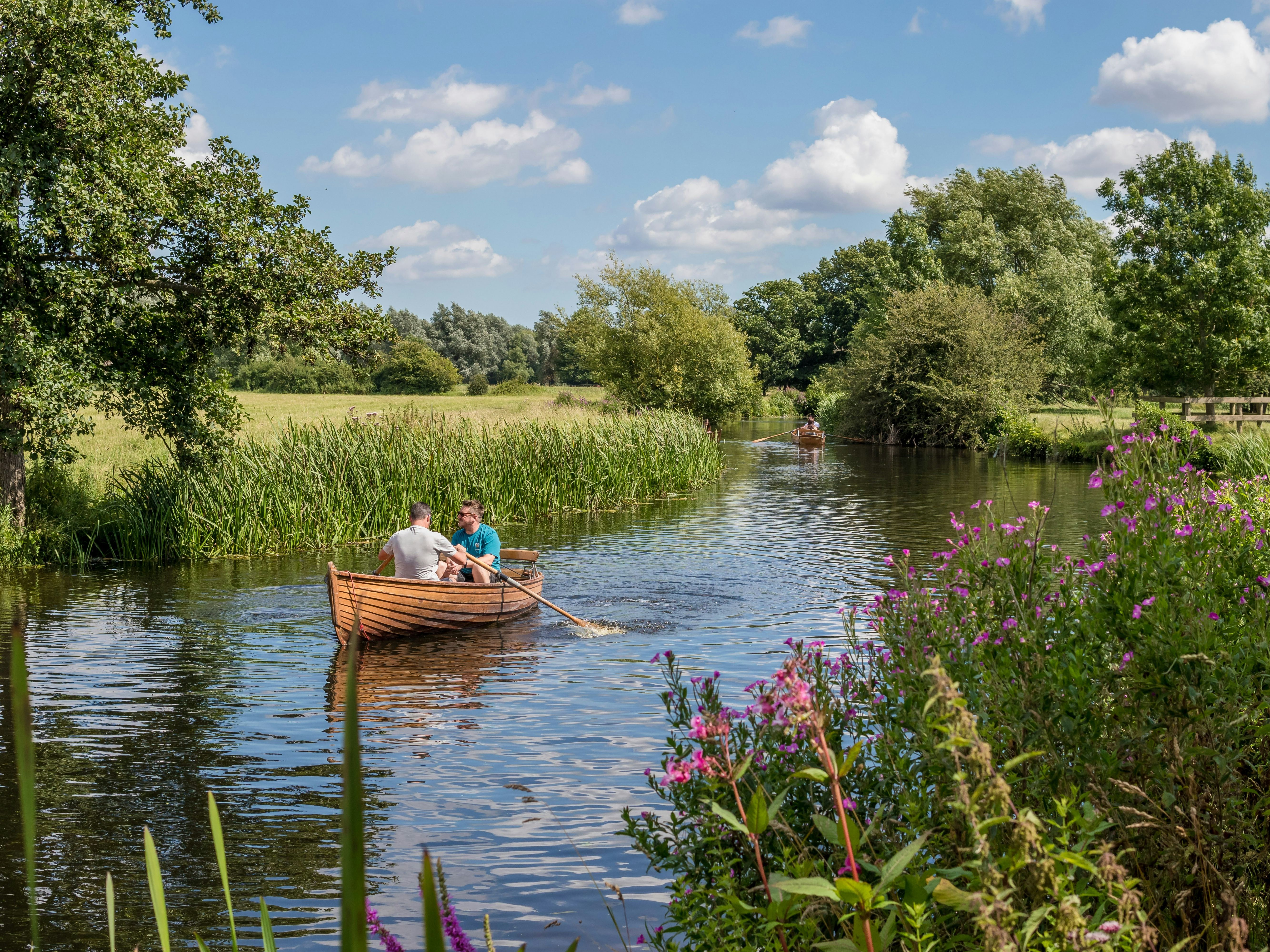 Two people rowing a wooden boat on a calm river surrounded by greenery and wildflowers.
